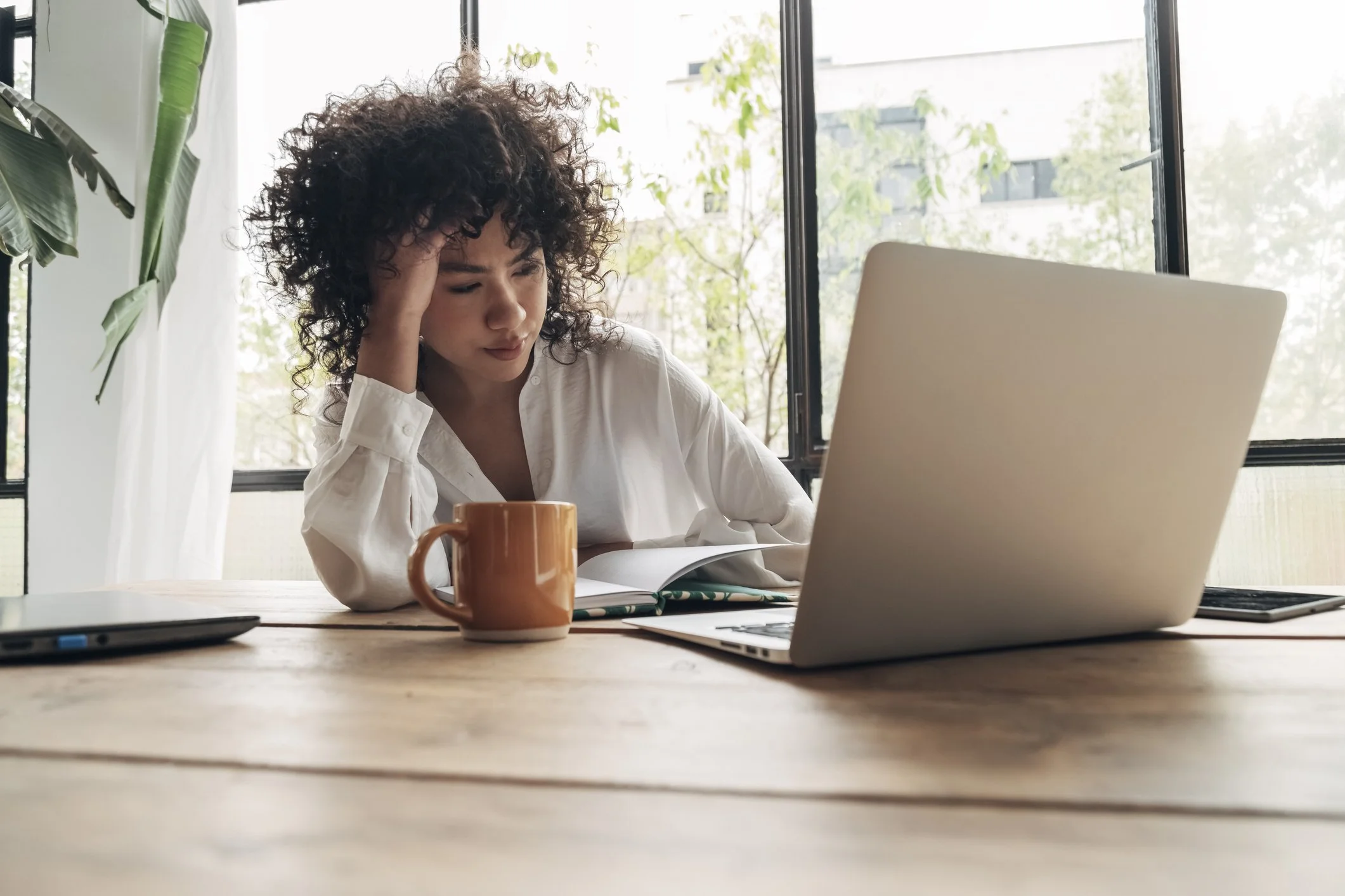 Woman with hand to head taking a mindful pause at her desk surrounded by plants and natural light, with laptop and coffee mug nearby