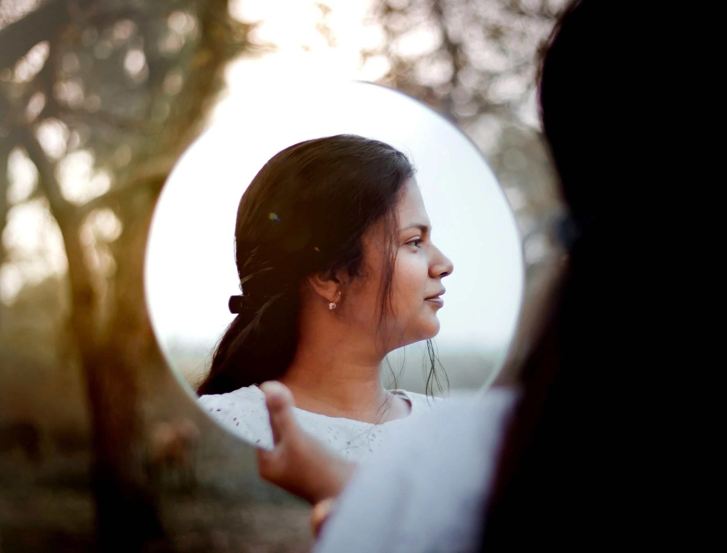 A woman looking at her reflection in a round mirror outdoors, representing reconnecting with yourself and rebuilding self-trust.