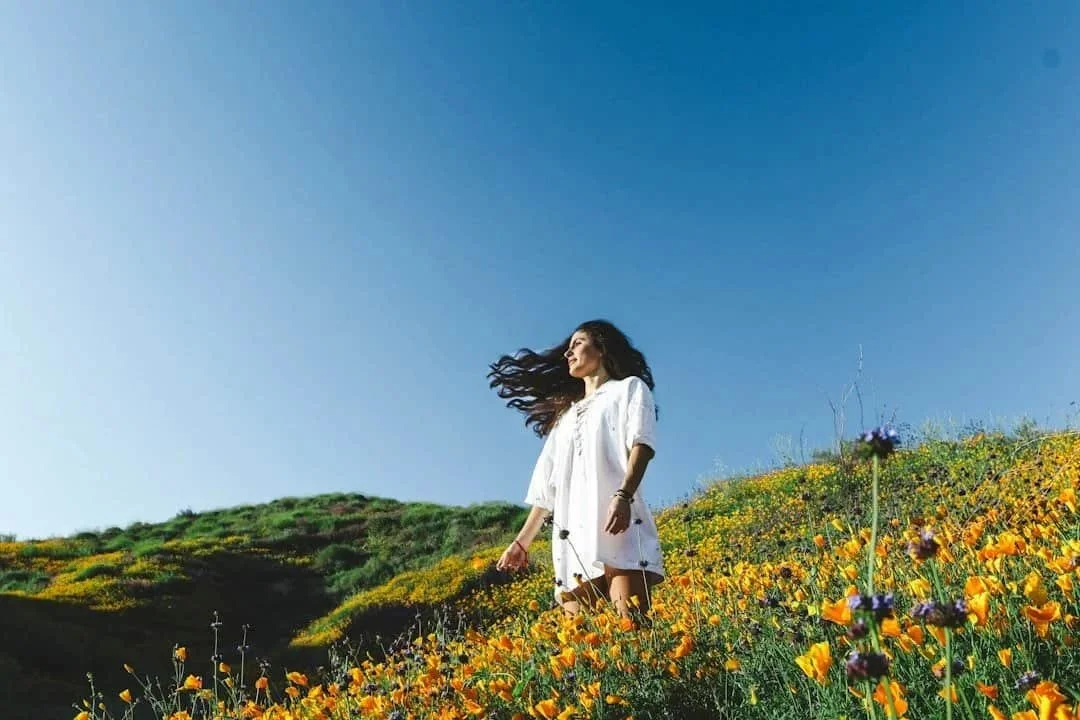 Woman in a white dress walking through a field of yellow wildflowers beneath a clear blue sky.