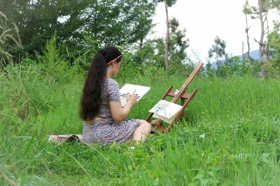Woman sitting in a grassy field painting at an easel surrounded by trees and greenery.