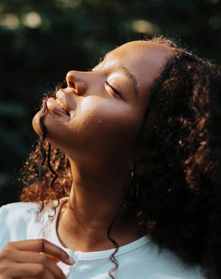 Young woman with curly hair standing outdoors with her eyes closed in the sunlight.