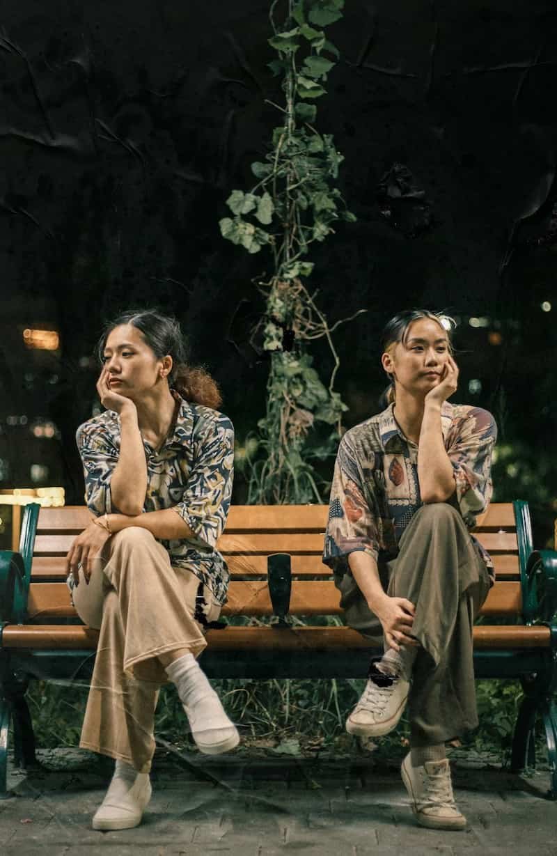 Two women sitting apart on a park bench, looking in opposite directions with thoughtful expressions.