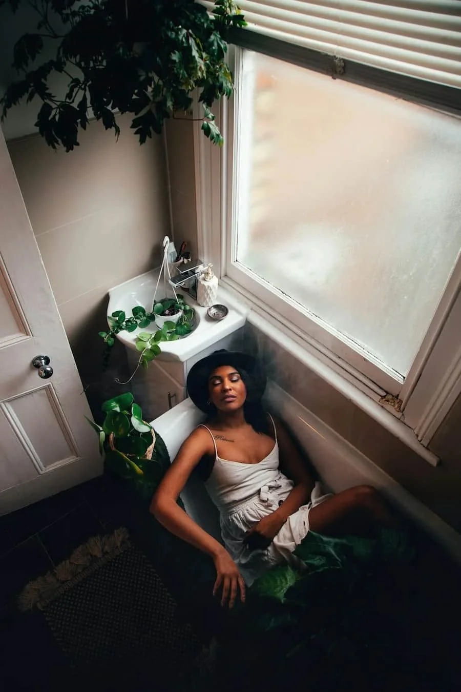 Woman resting in a bathtub beside a large window and houseplants.