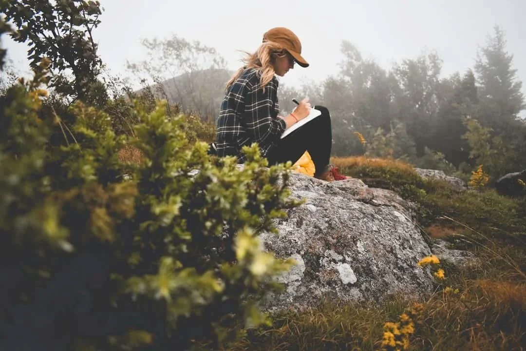 Young adult journaling on a rocky hillside in nature, reflecting on life transitions and personal growth.