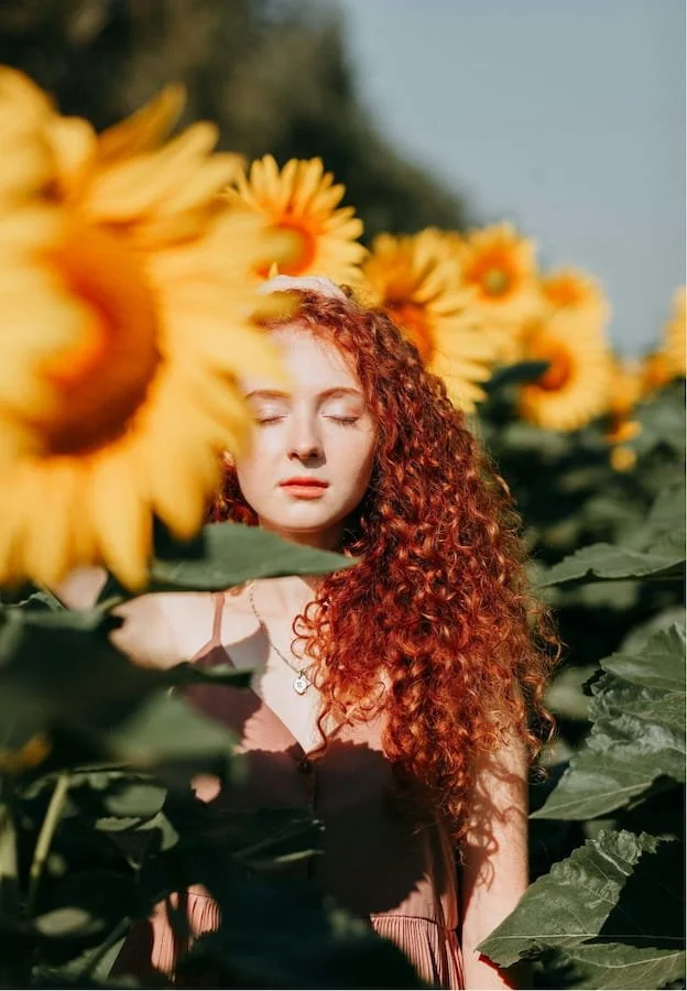 Woman with curly red hair standing among yellow flowers with her eyes closed.
