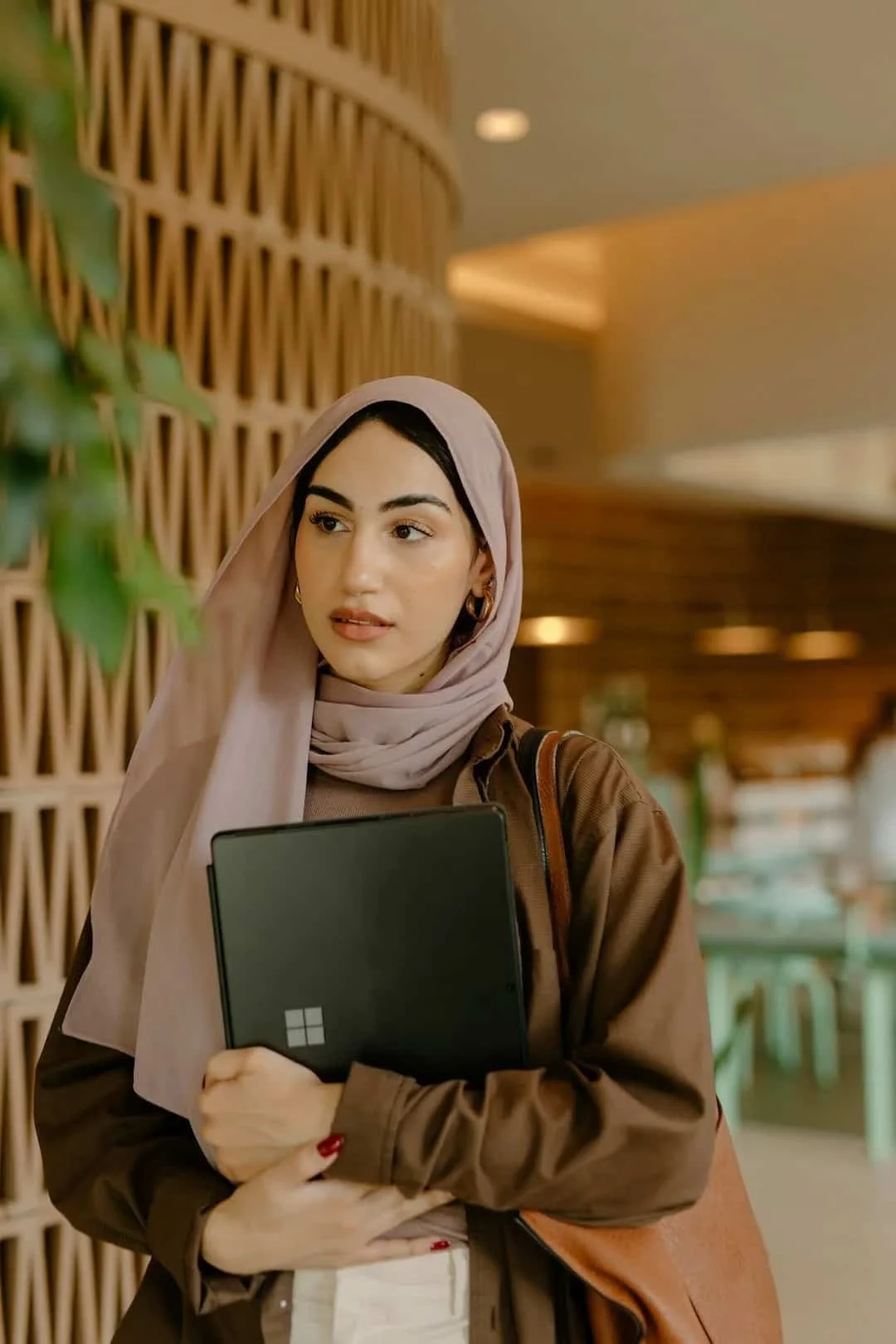 Woman wearing a light-colored hijab and holding a laptop while standing indoors.