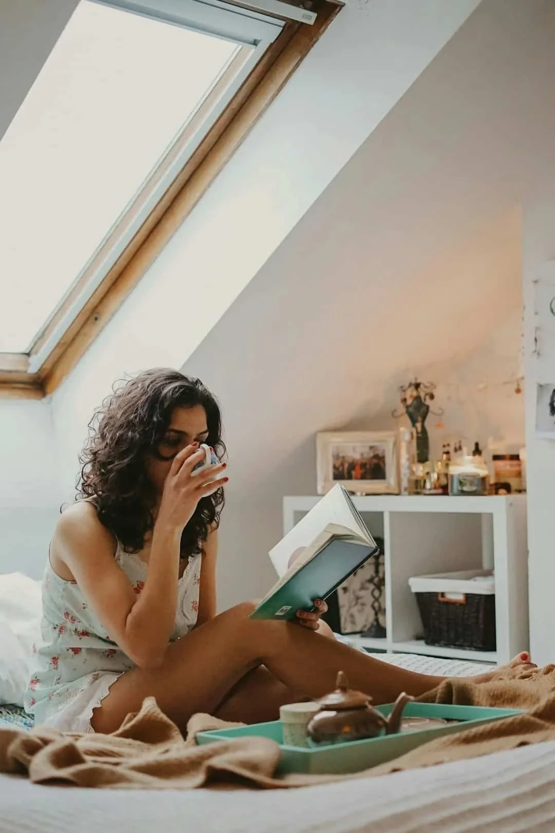 Woman sitting on a bed reading and drinking tea in a calm morning space, representing reflection and nervous system regulation for anxiety.