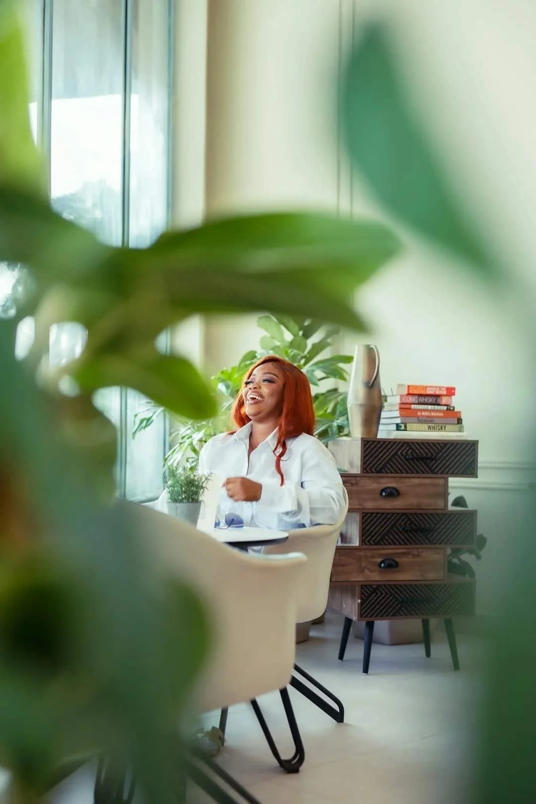 Woman laughing while sitting in a bright room surrounded by plants.