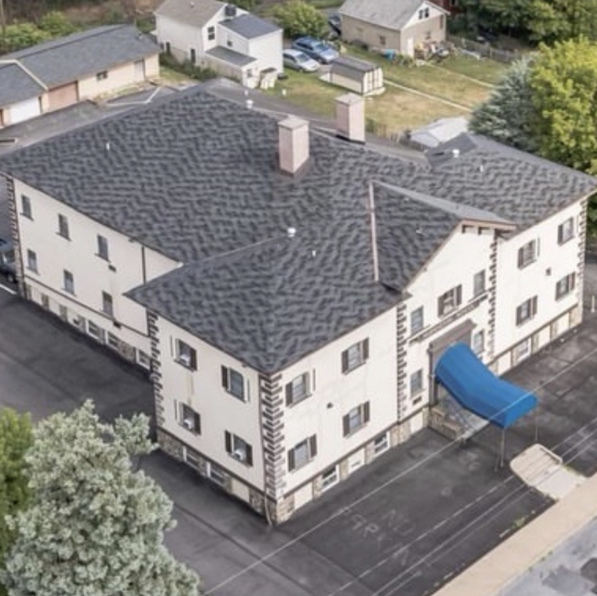 Aerial view of a large three-story building with a dark patterned roof and a blue awning over the entrance. The building is surrounded by a paved parking area and nearby residential houses.