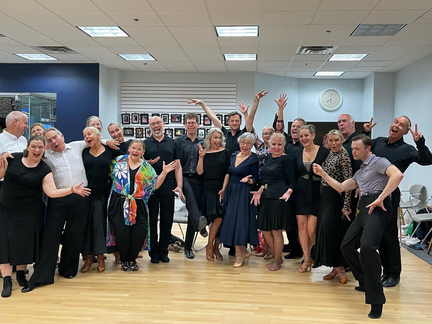Group of people celebrating happily in a room with a wooden floor, some with arms raised and smiling, in festive or dance attire.
