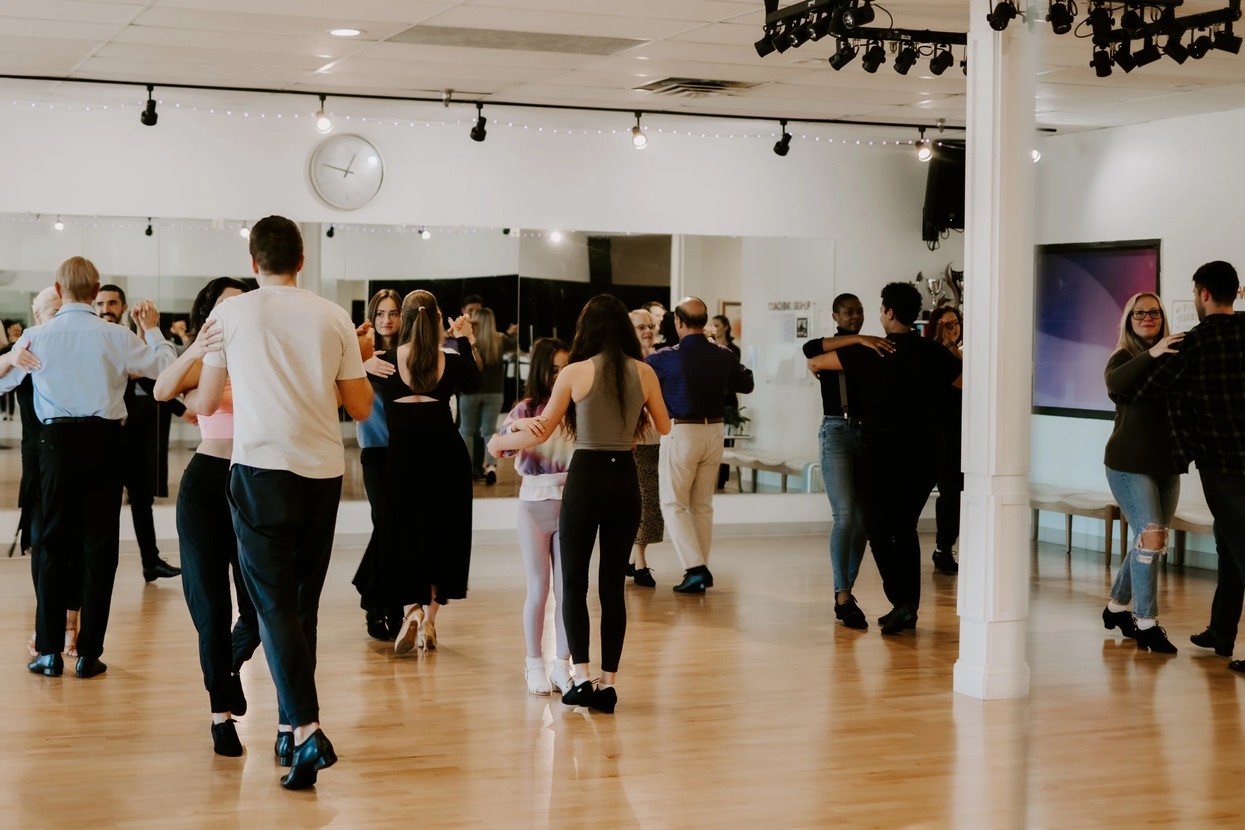People dancing in a dance studio with wood floors, white walls, a large mirror, and a clock on the wall.