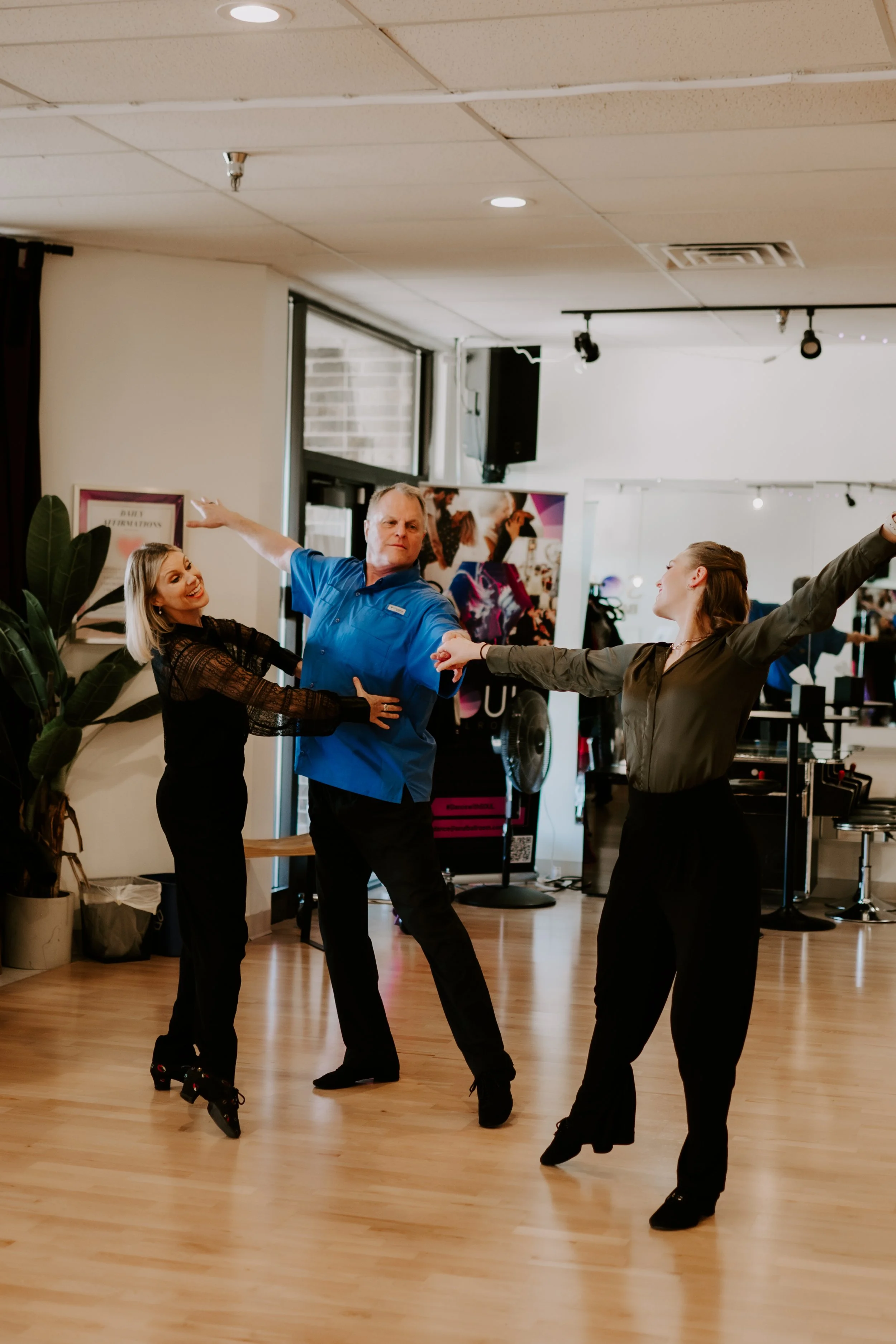 Three people dancing in a studio, with two women wearing black and a man in a blue shirt. They are holding hands and smiling, with a woman in black's arms outstretched.