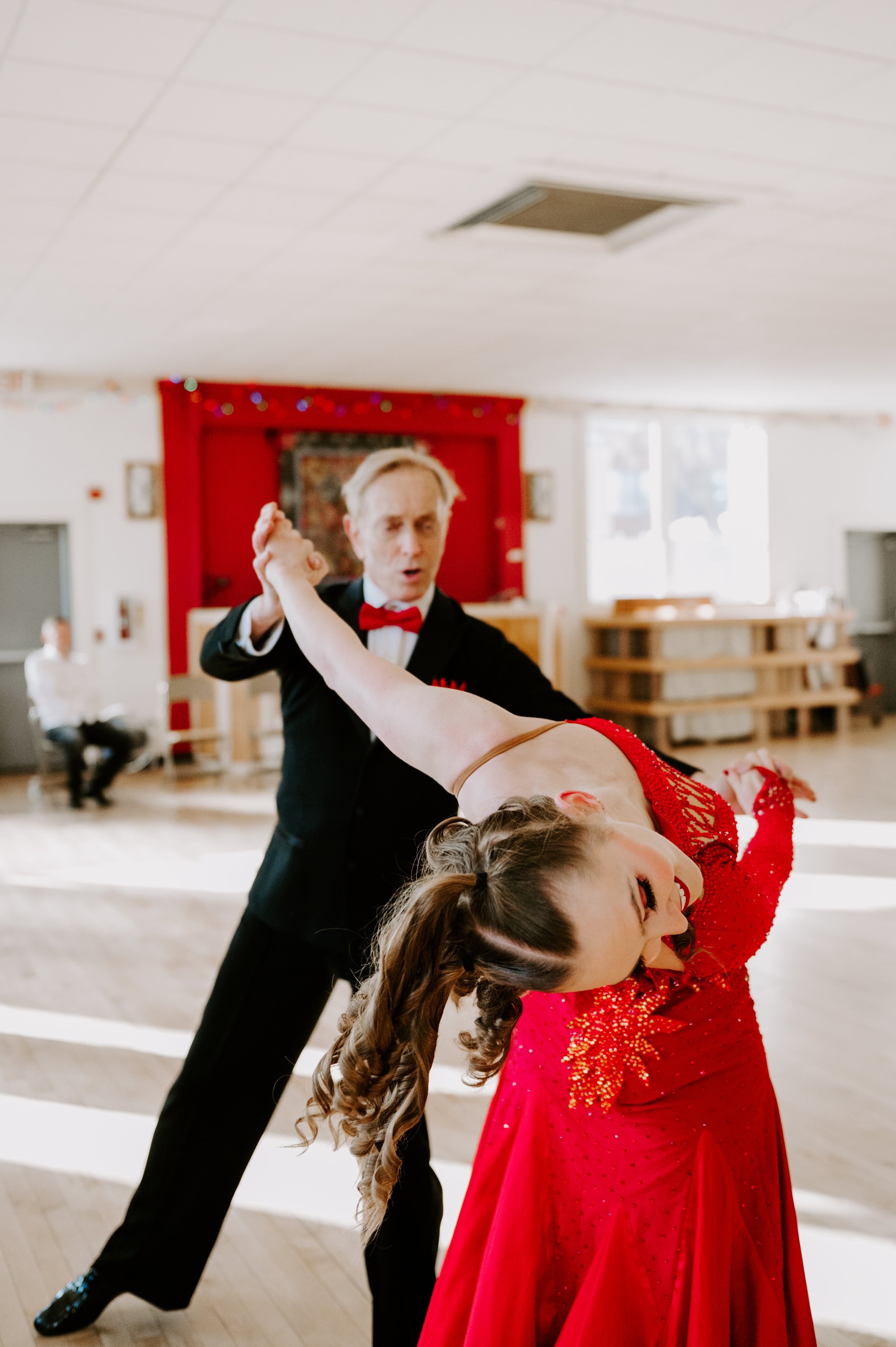 A man and woman are dancing, with the woman leaning back in a dip. The man, wearing a black tuxedo and red bow tie, looks surprised while holding the woman's hand. The woman, in a bright red, sparkling dress with flowing hair, is smiling. The background shows a decorated indoor space with a red wall, windows, and a person sitting in the background.