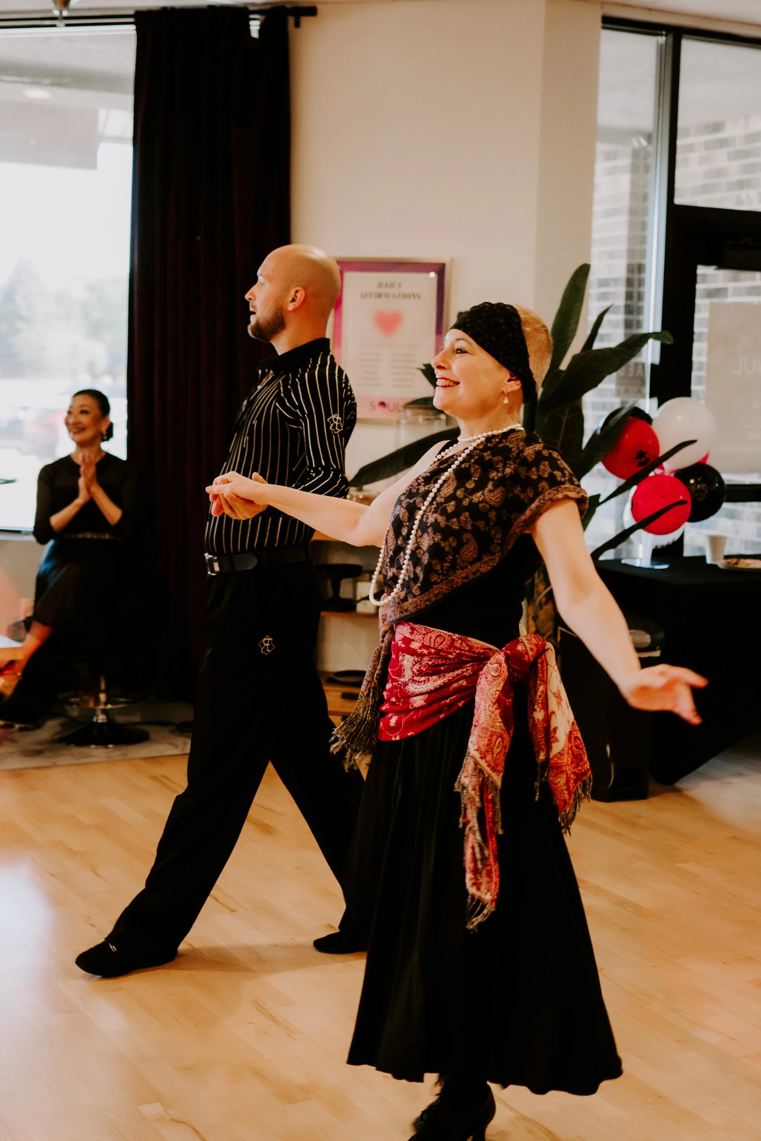Women and a man dancing in a room with wooden floor, decorated for a celebration with balloons and a large plant.