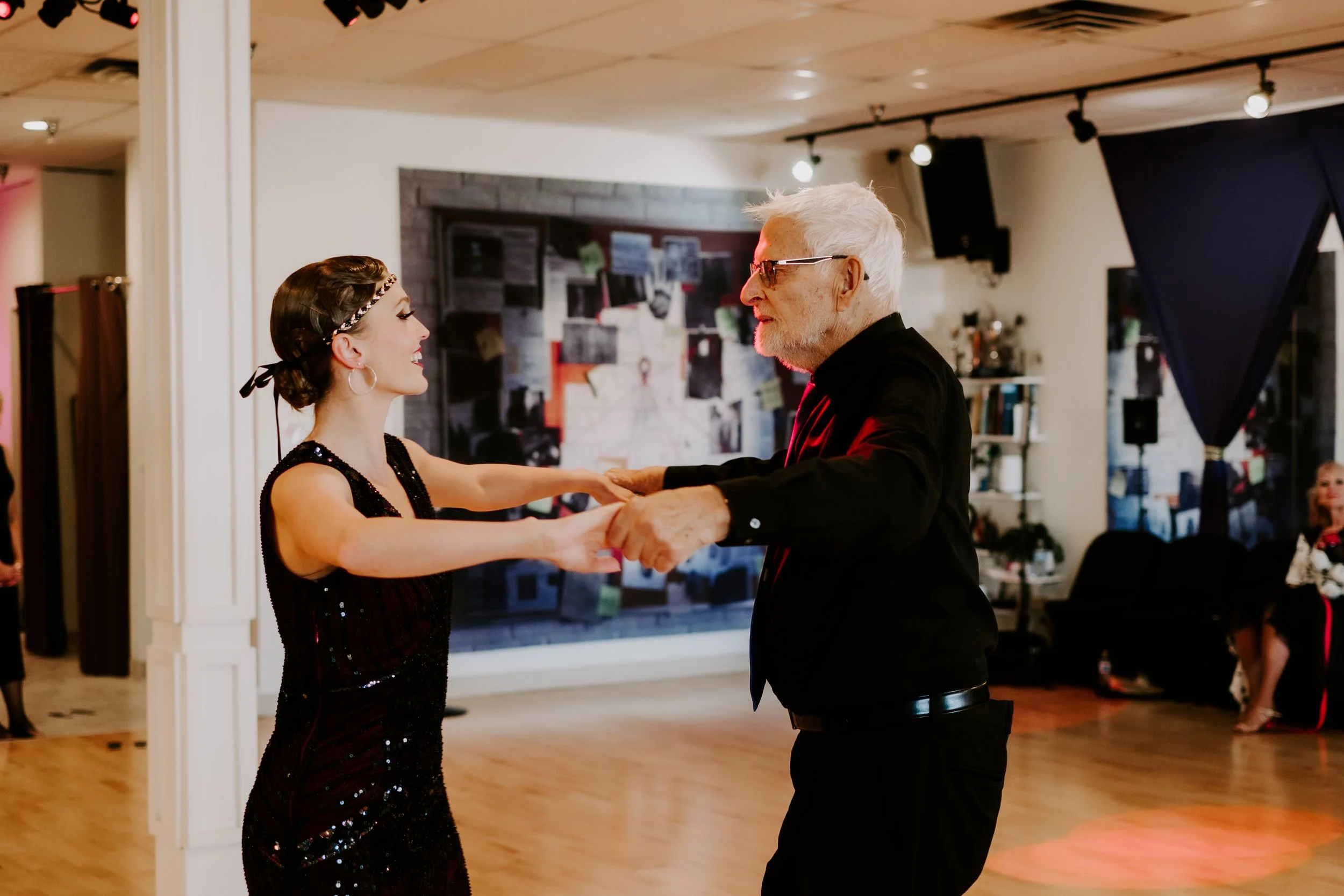 Older man and young woman dancing together in a dance studio, smiling and holding hands.