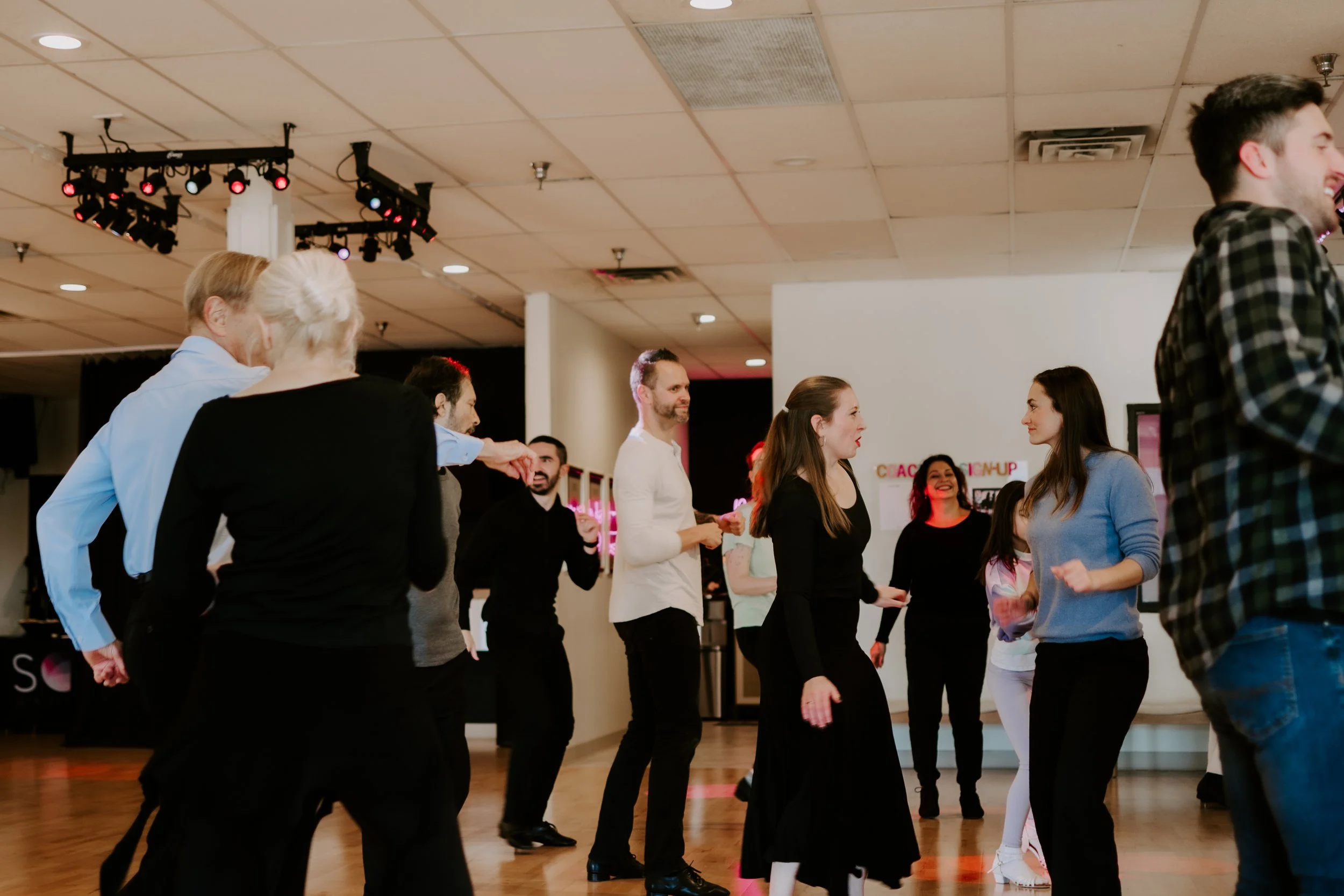 People dancing and socializing in a dance studio with a wooden floor, ceiling lights, and stage lighting equipment.