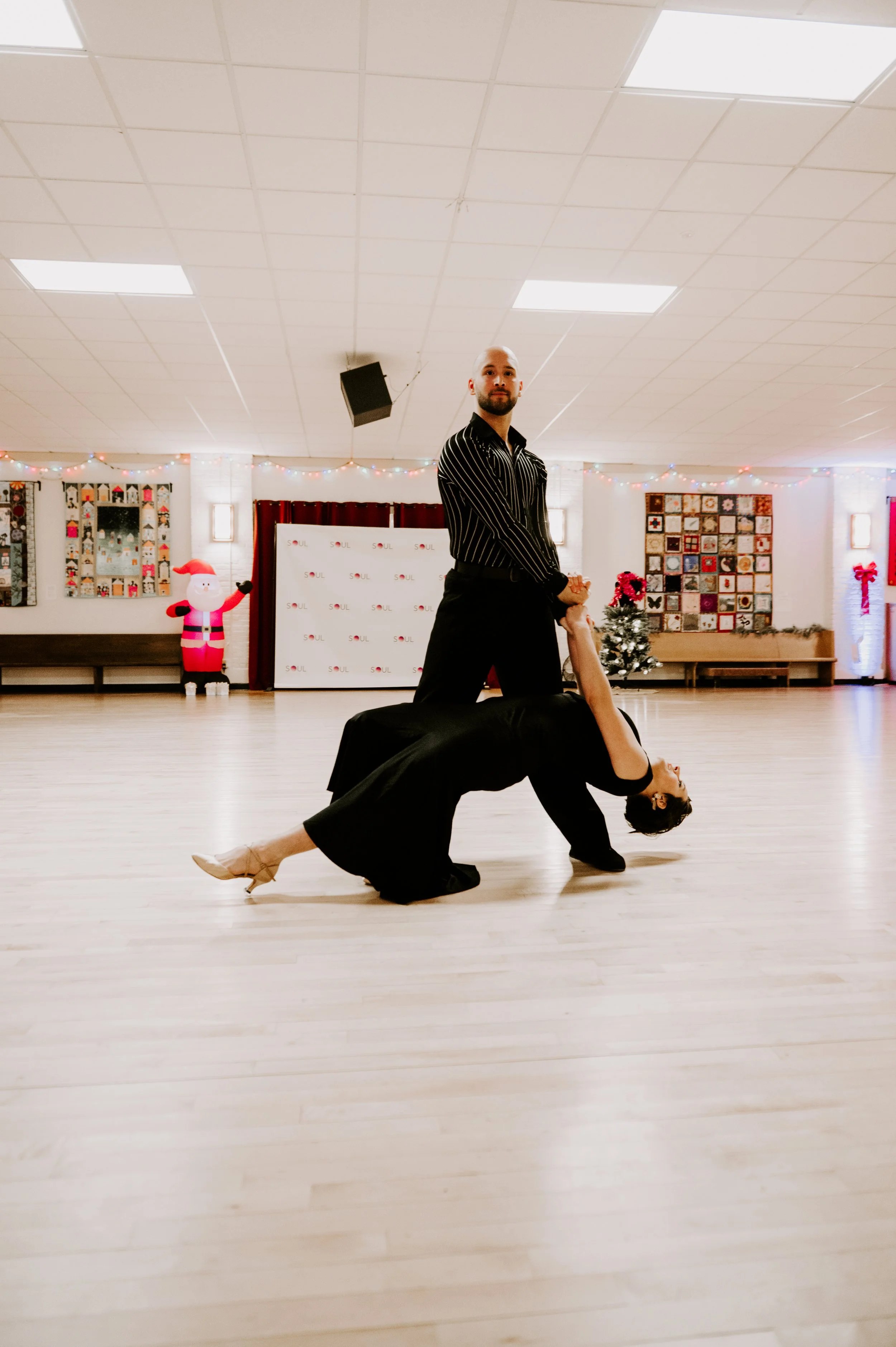 A man and woman dance together in a spacious room decorated for Christmas, with a Christmas tree, holiday lights, and holiday-themed decorations in the background.