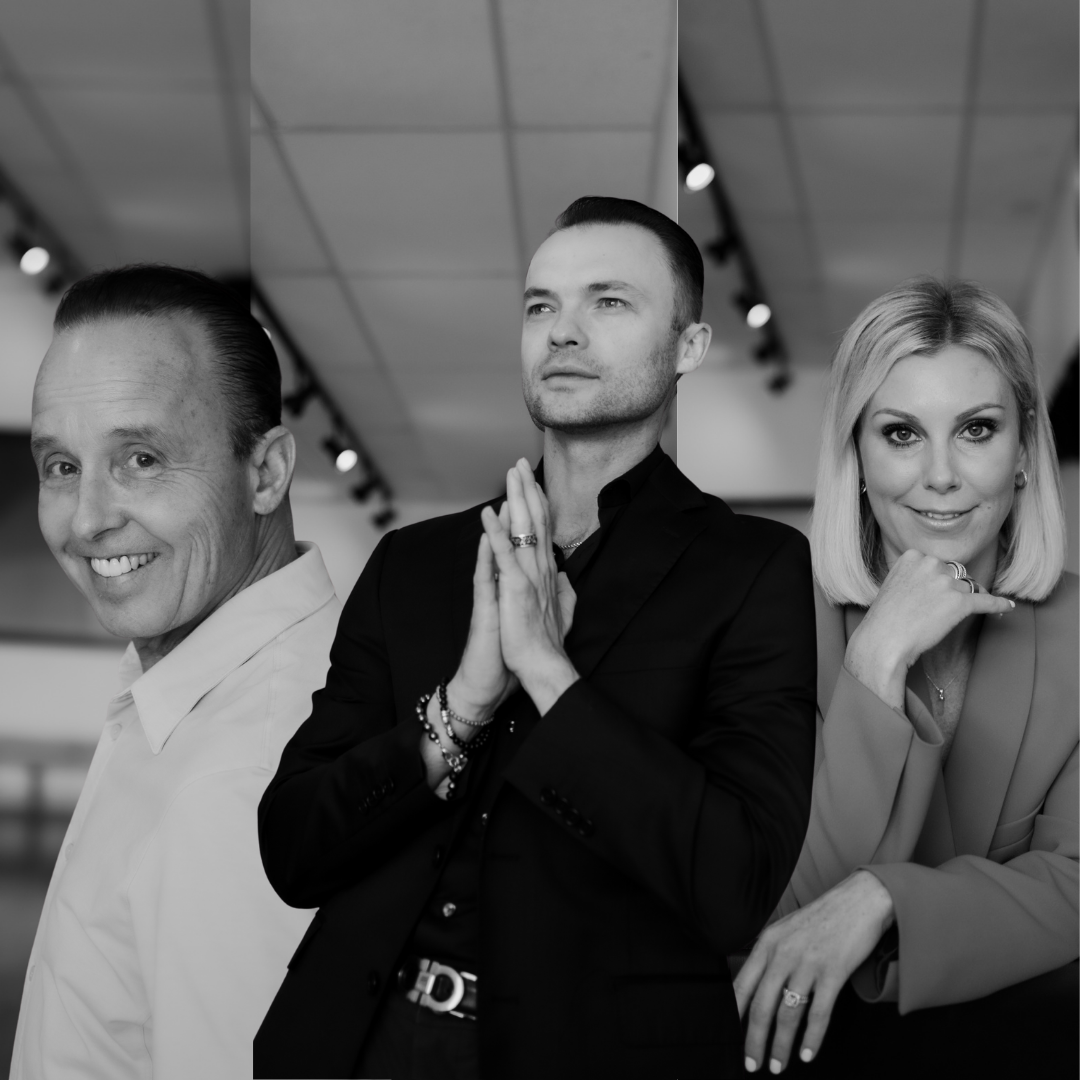 Black and white photo of three adults standing indoors, with the man in the middle having hands pressed together in prayer, and a woman on the right resting her chin on her hand, all looking confidently towards the camera.