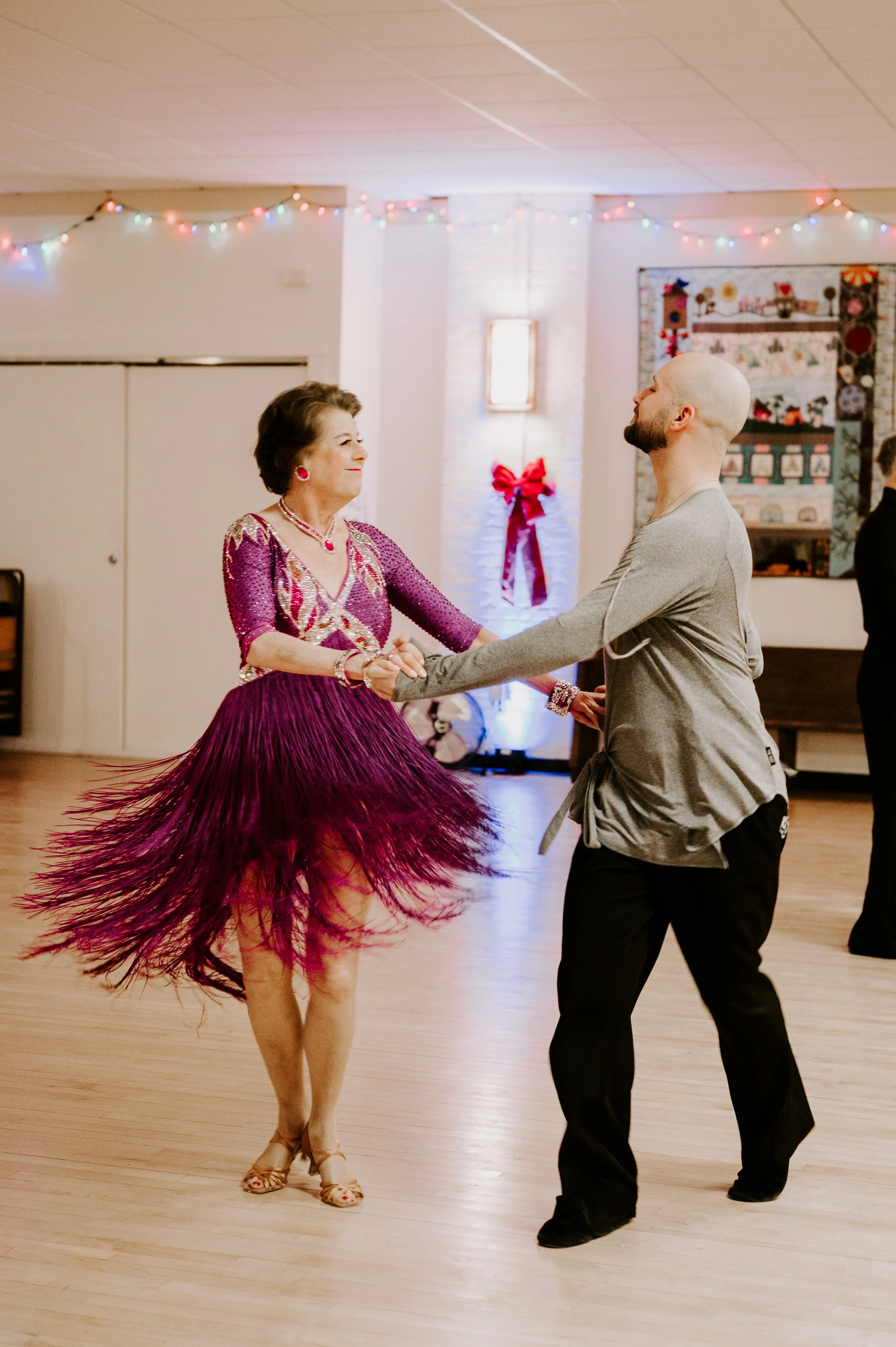 A woman and a man are dancing together in a decorated room, with the woman wearing a purple dress with a fringe skirt and the man in a gray hoodie and black pants.