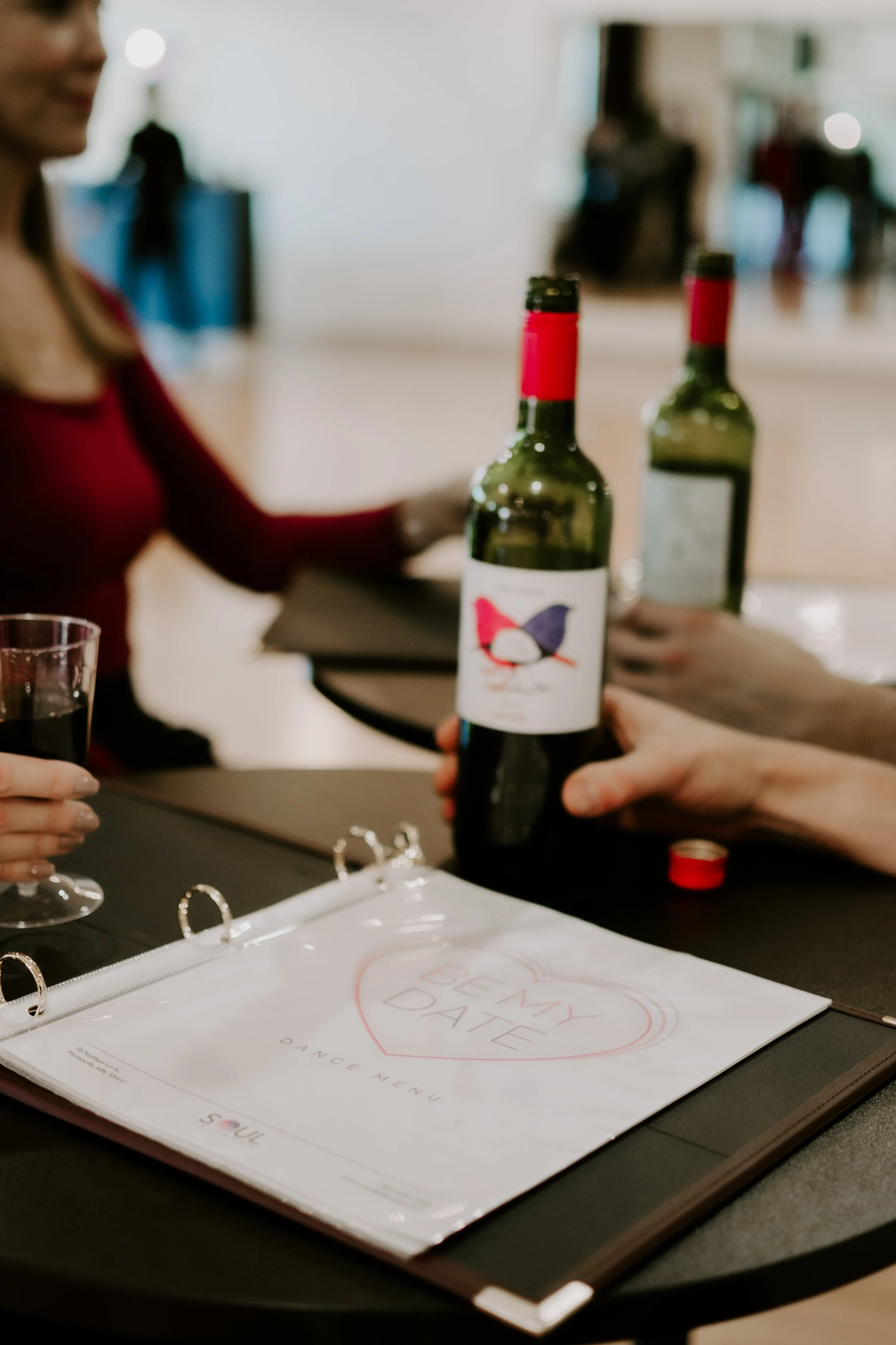 A single red rose in a glass bottle on a table, with blurred people in the background.