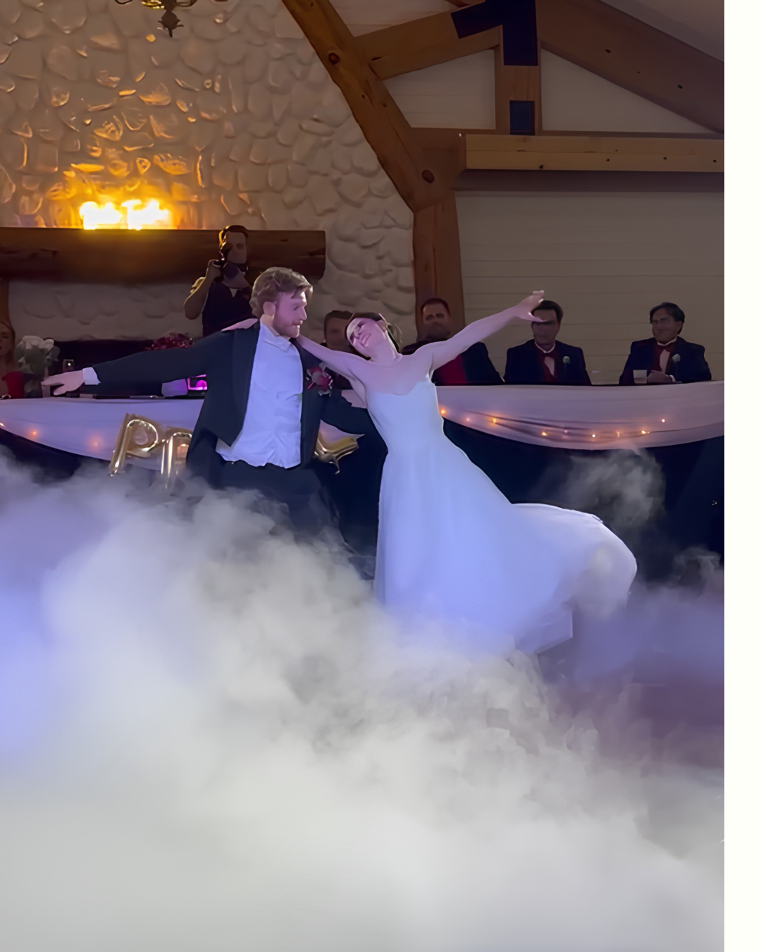 A bride and groom dancing at their wedding reception, surrounded by fog and dim lighting, with guests seated in the background.