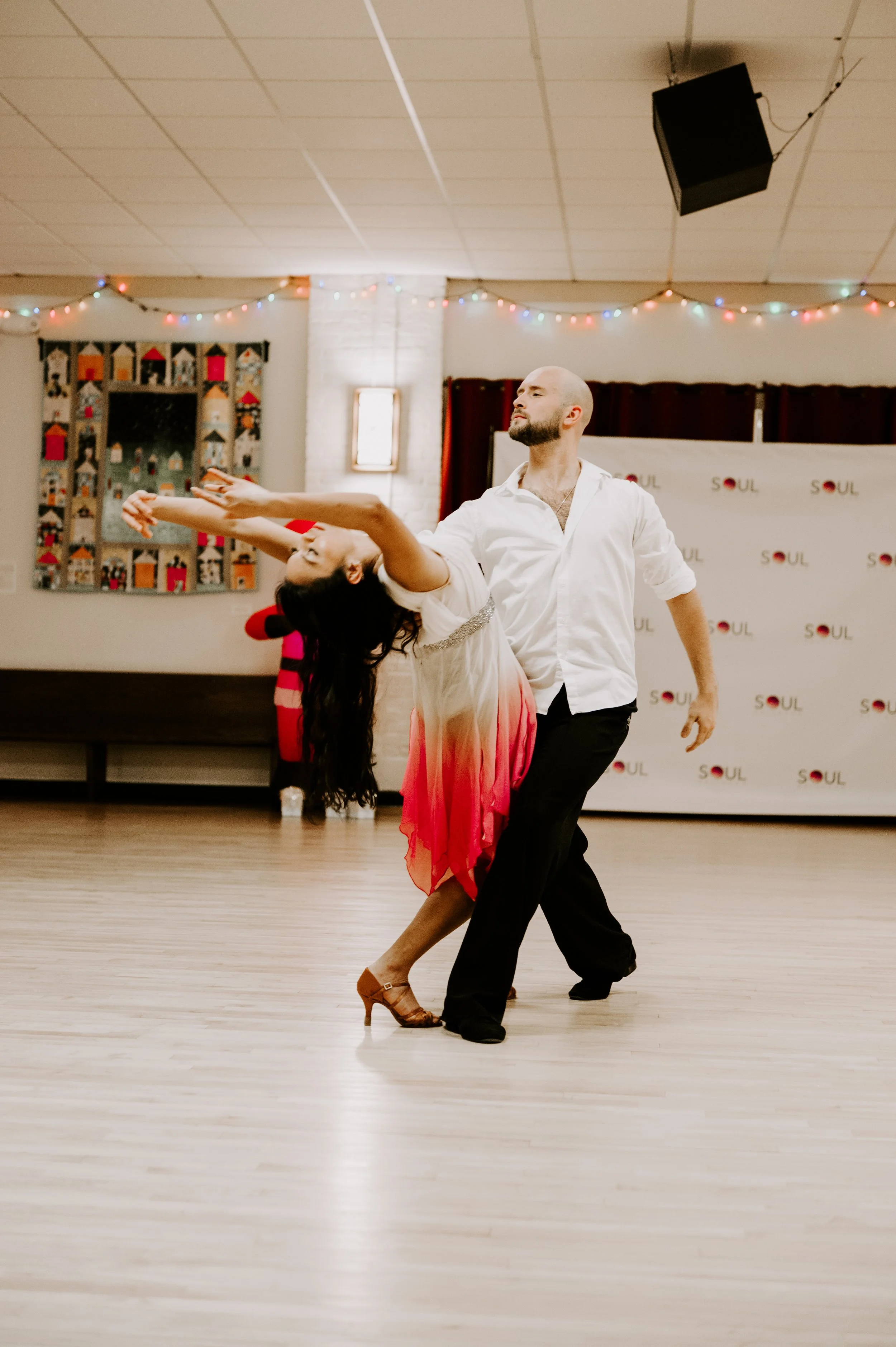 A man and woman dancing together in a room with wooden flooring and festive string lights, with a backdrop featuring the word 'SOUL' repeated.