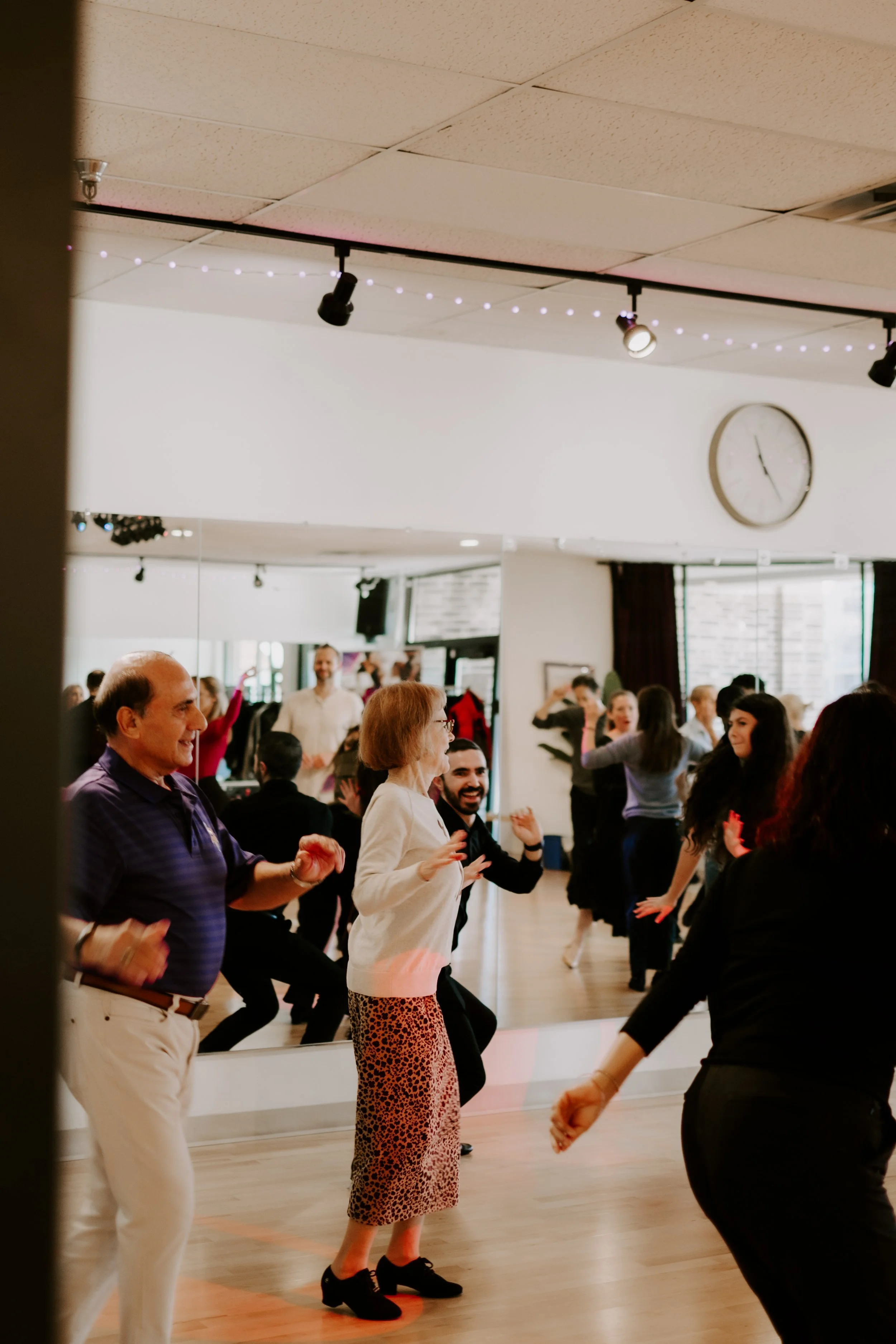 People dancing happily in a dance studio with a large mirror, clock, and ceiling lights.