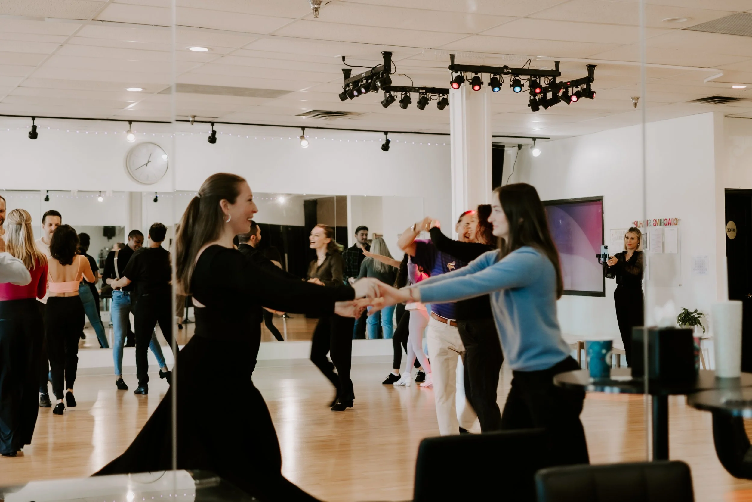People dancing in a dance studio, with some holding hands and smiling, surrounded by mirrors and dance studio equipment.