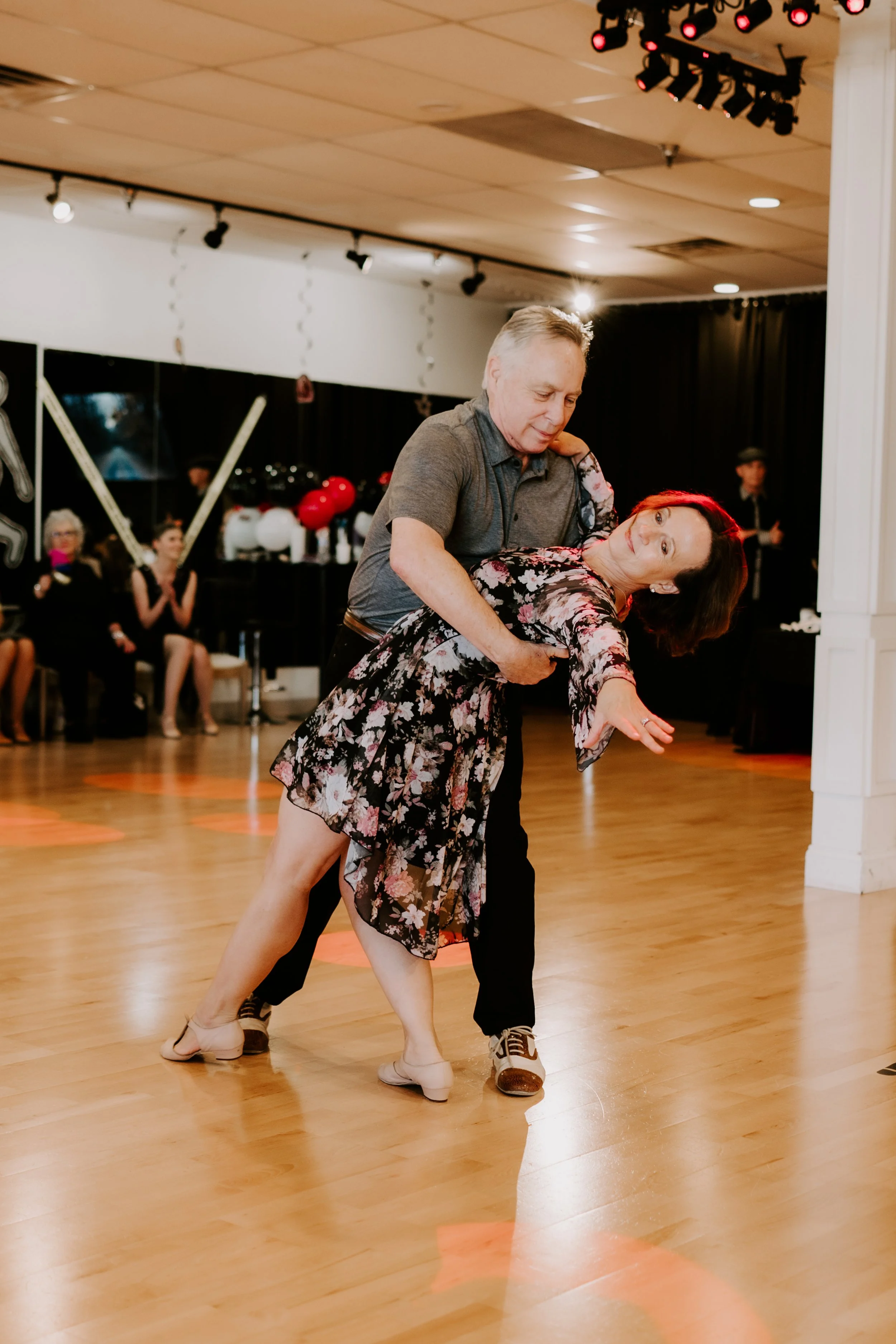 A man dancing with a woman in a floral dress at a social gathering or dance event, with other people seated and clapping in the background.