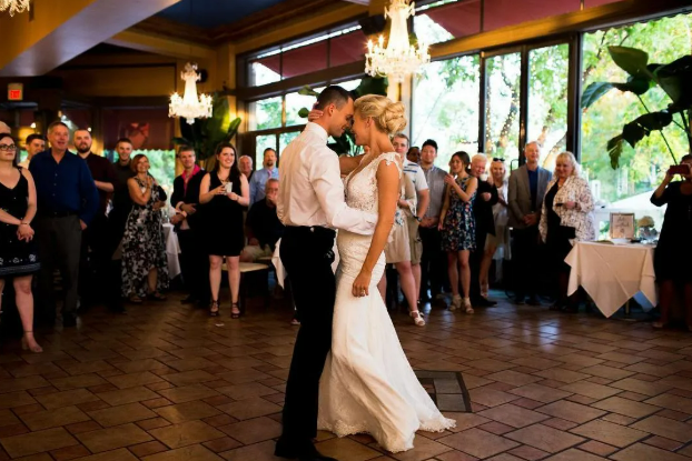 A bride and groom dancing closely at their wedding reception, surrounded by guests watching them in a warmly decorated venue with chandeliers and large windows.