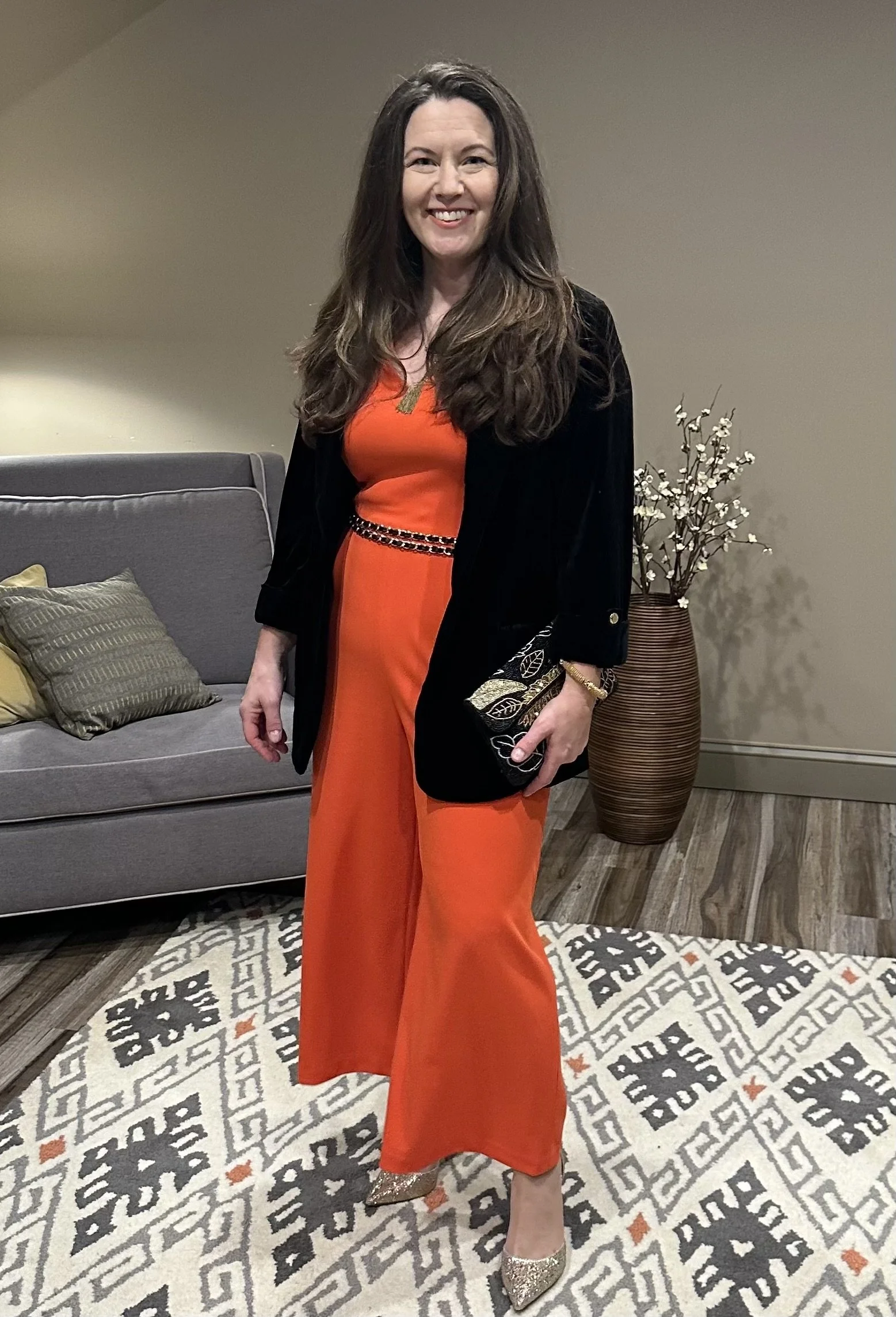 Woman in an orange jumpsuit and black velvet jacket standing in a living room with a gray sofa, decorative pillows, and a tall vase with white flowers.