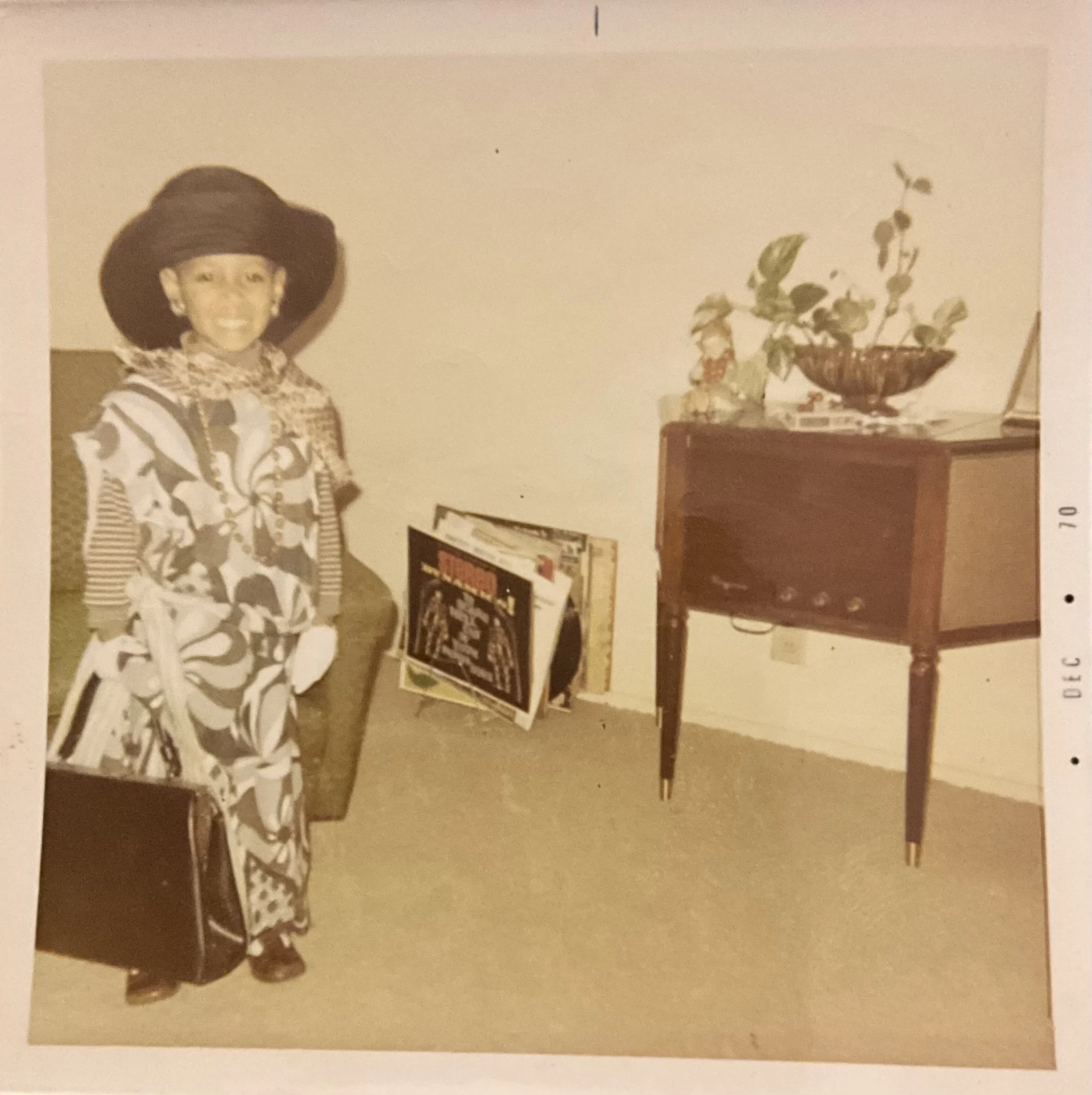A small child dressed in an oversized floral dress, wearing a large black hat, standing next to a chair with a handbag. There are vinyl records leaning against the wall, and a wooden table with a plant in a bowl and some decorative items on it.