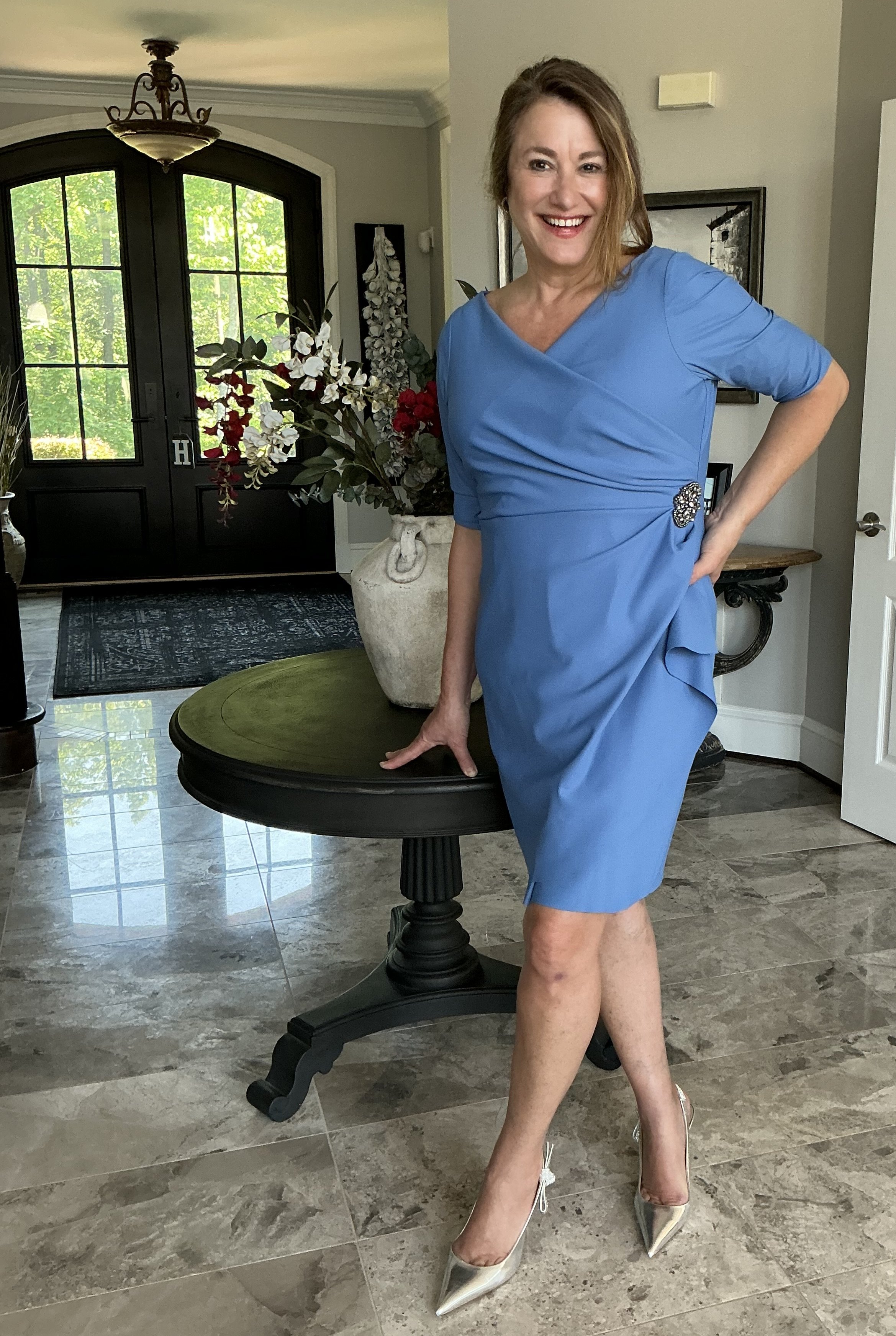 A woman in a blue dress standing beside a round table with a large white vase of flowers inside. She has one hand on the table and is smiling at the camera. The background features a front door with glass panels, a chandelier, and decorative wall art