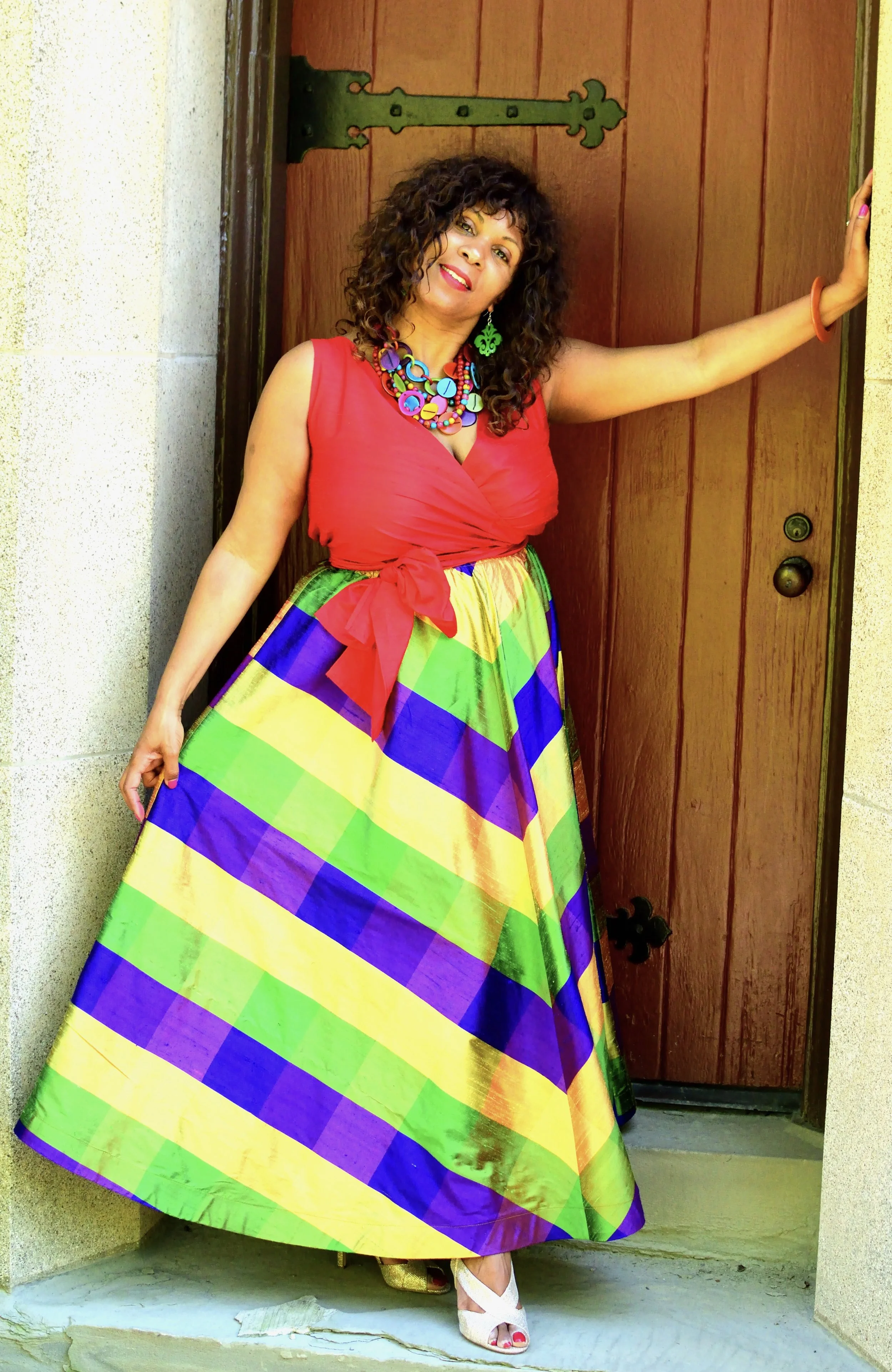 A woman with curly dark hair standing by a wooden door, wearing a colorful dress with a rainbow plaid pattern, red top, and accessorized with multicolored jewelry and a bracelet.