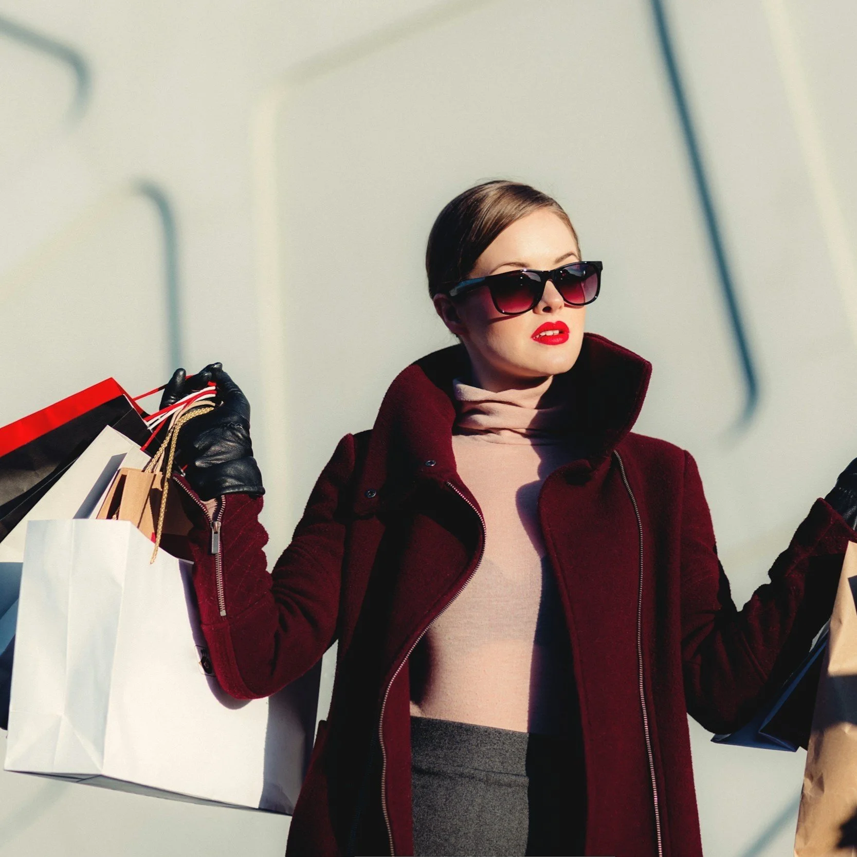 A woman with short brown hair wearing large black sunglasses, a beige turtleneck, a dark red coat, and black gloves, holding shopping bags in both hands.