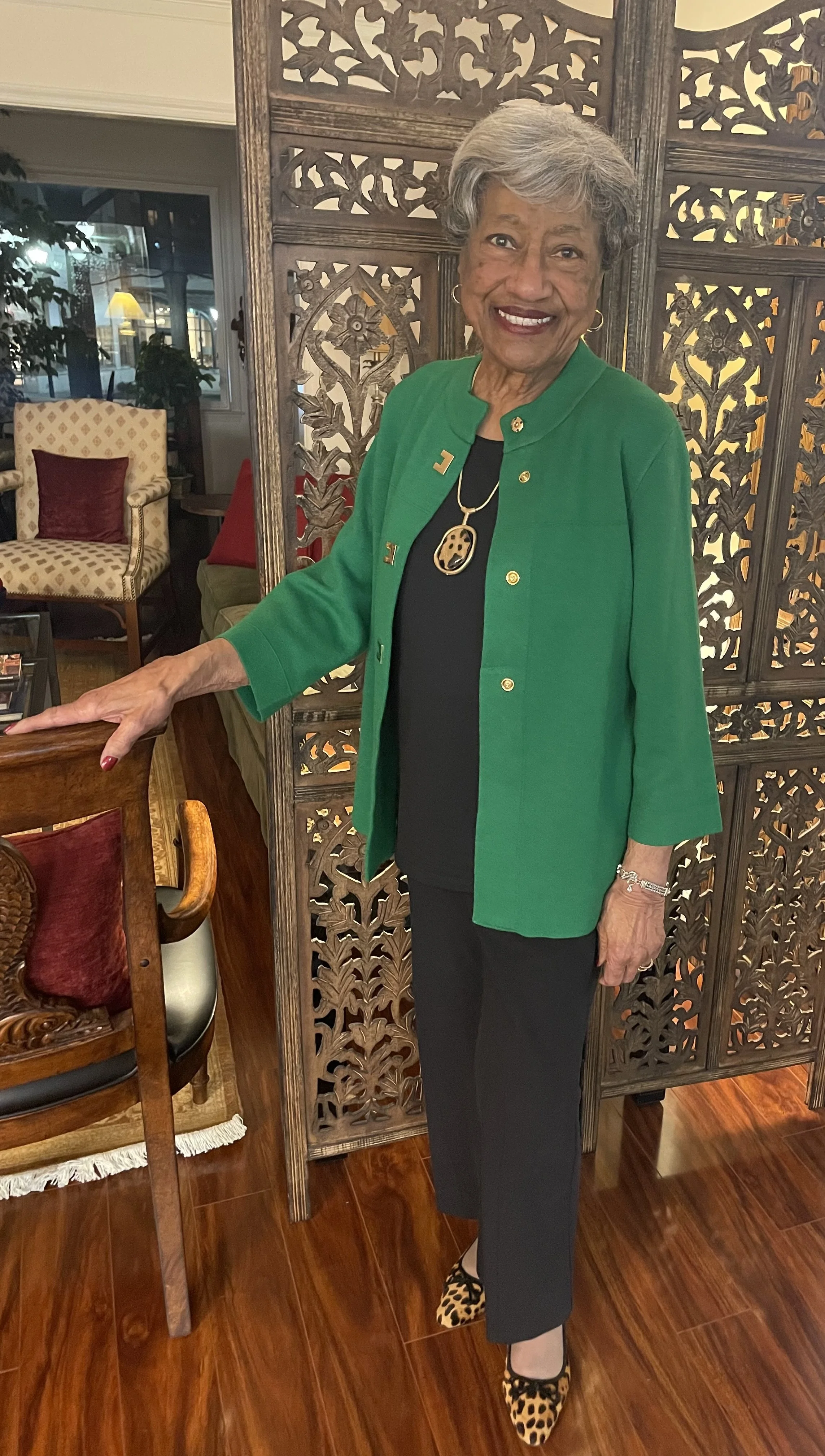 An elderly woman with gray hair smiling, wearing a green jacket, black top, black pants, leopard print shoes, and jewelry, standing inside a room with wooden furniture and decorative wooden room divider.