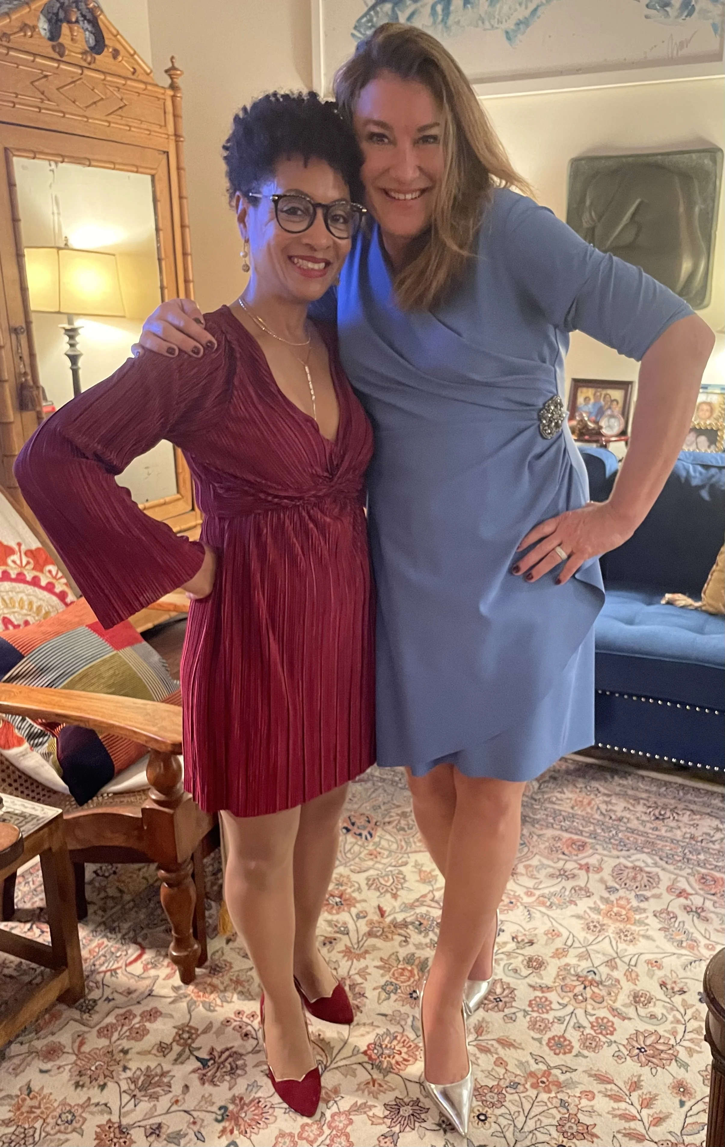 Two women standing together in a living room, smiling. One woman is wearing a red dress with flared sleeves and red shoes, and the other is wearing a light blue dress with metallic silver shoes. They are posing with their arms around each other.