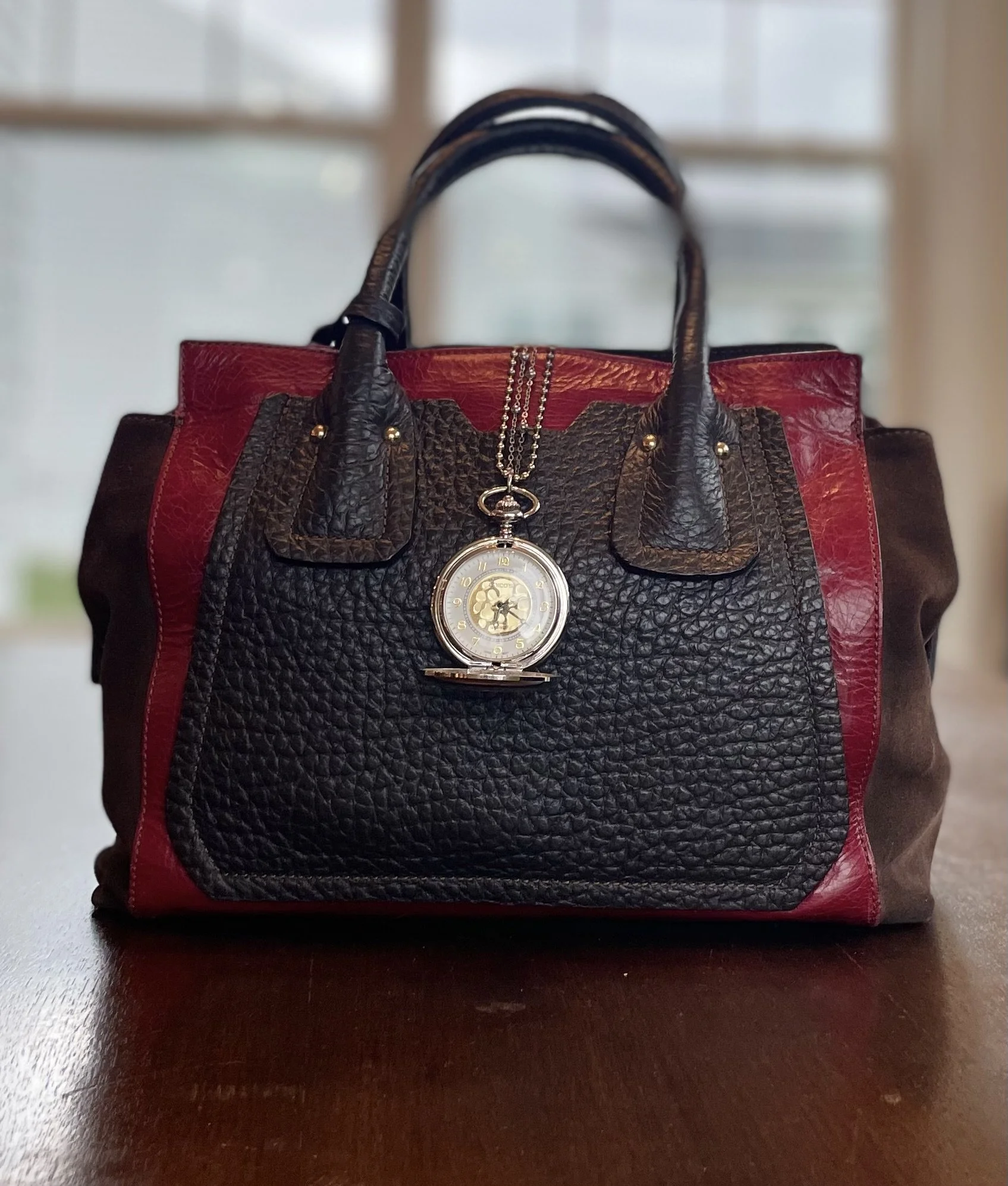 A handbag with red, black, and brown leather and a silver pocket watch hanging from the handle.