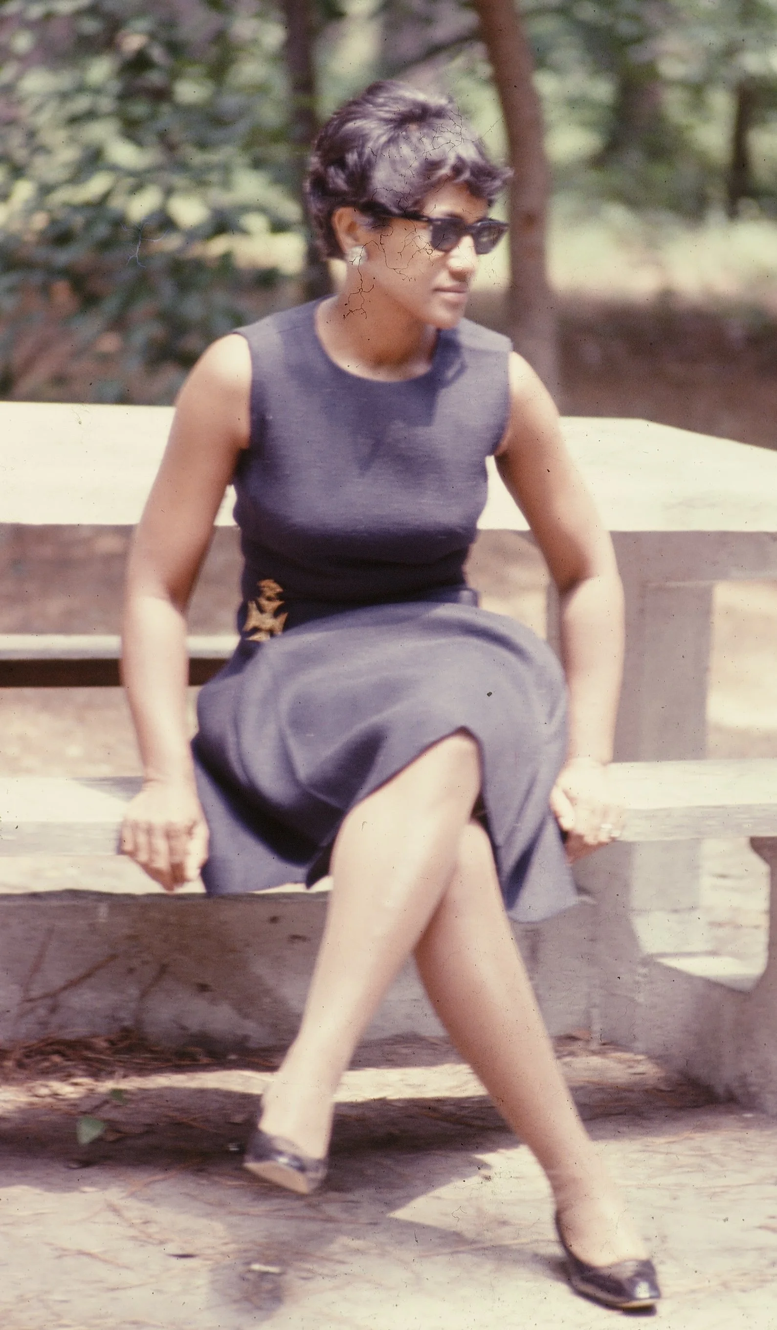 A woman sitting on a park bench, wearing sunglasses and a sleeveless black top with a long dark skirt, with her legs crossed, in an outdoor setting with trees in the background.