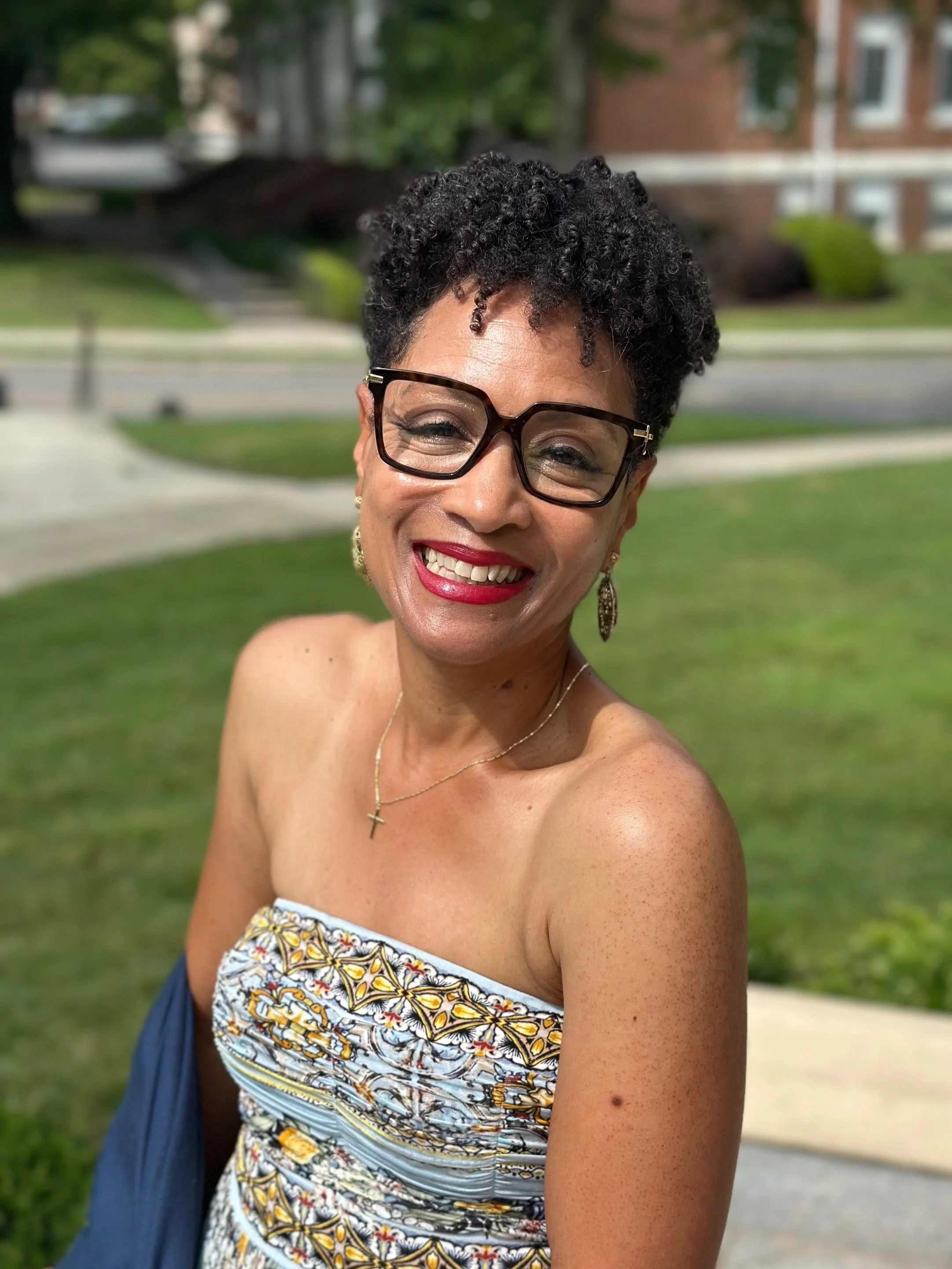 A smiling woman with short curly black hair, wearing glasses, red lipstick, a patterned strapless top, earrings, and a necklace with a cross, sitting outdoors on a park bench with a grassy area and residential buildings in the background.