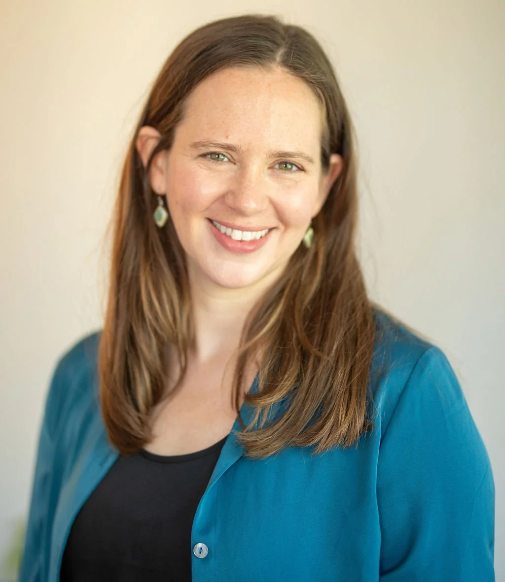 Smiling woman with long brown hair wearing a teal jacket and black shirt, against a neutral background.