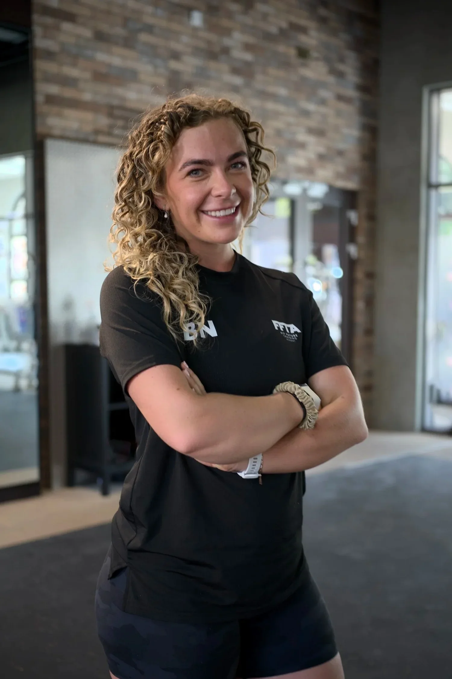 A woman with curly blonde hair smiling, standing with arms crossed inside a building with brick walls and large windows, wearing a black athletic shirt with logos and black shorts.