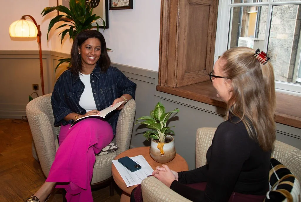 Two women sitting in chairs having a conversation in a cozy room, with a potted plant on a small table between them and a window in the background.