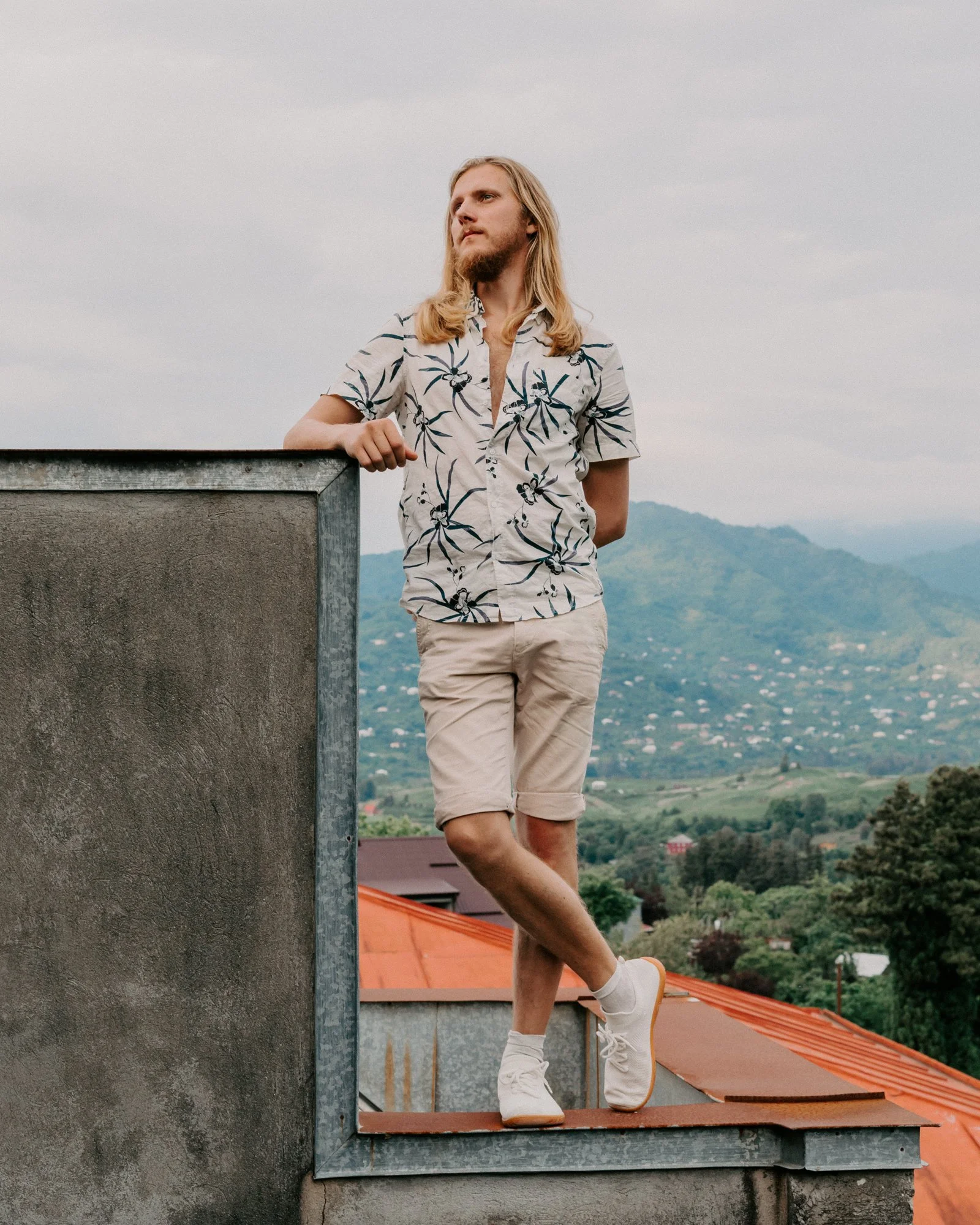 A young man with long blond hair and a beard, wearing a short-sleeved floral shirt and khaki shorts, is standing on a ledge next to a concrete wall. He is outdoors with a mountainous landscape and houses with red roofs in the background, looking thou