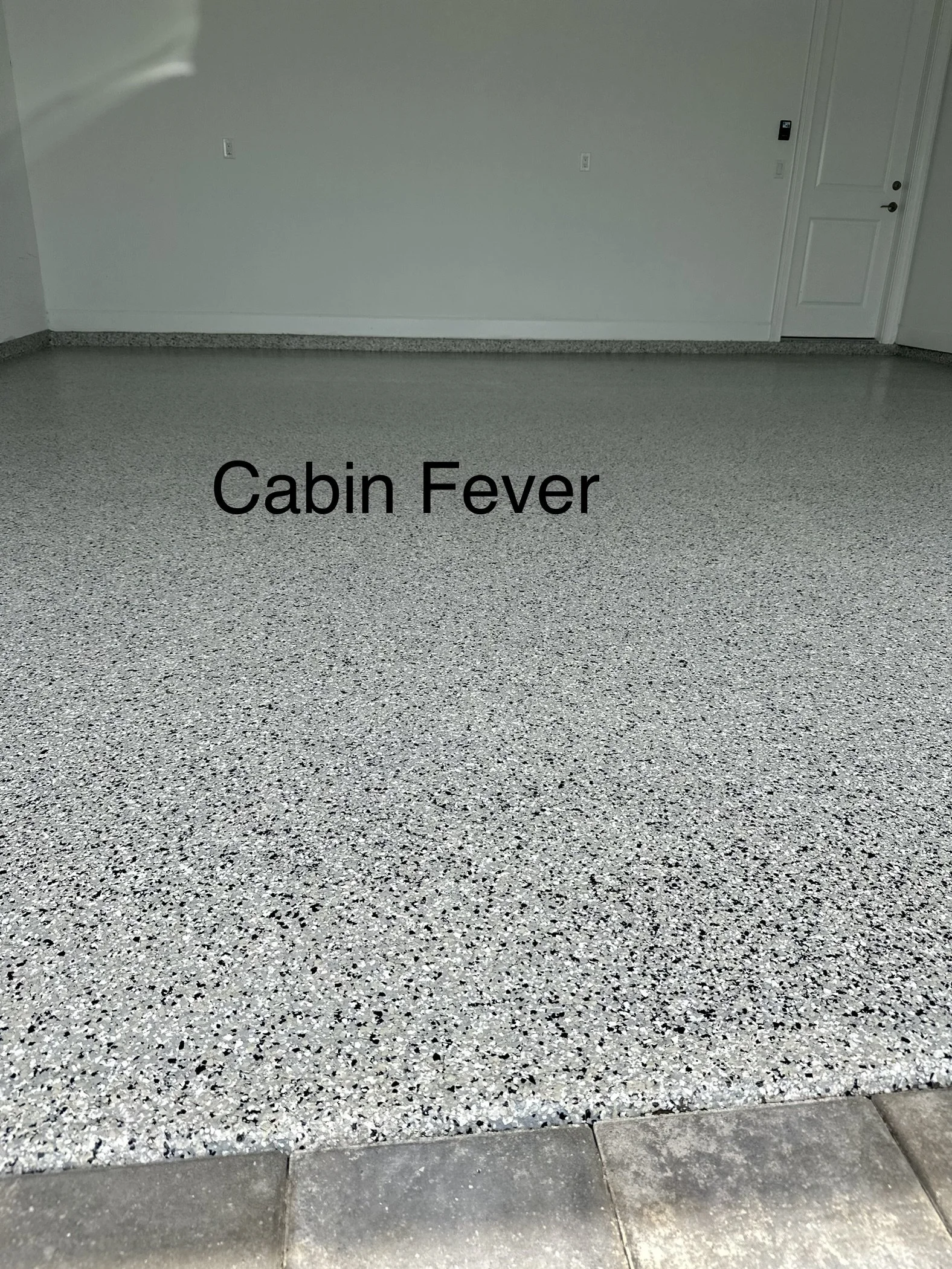 Empty room with a speckled epoxy floor, white walls, a closed white door, and electrical outlets. The text 'Cabin Fever' is overlaid on the floor.