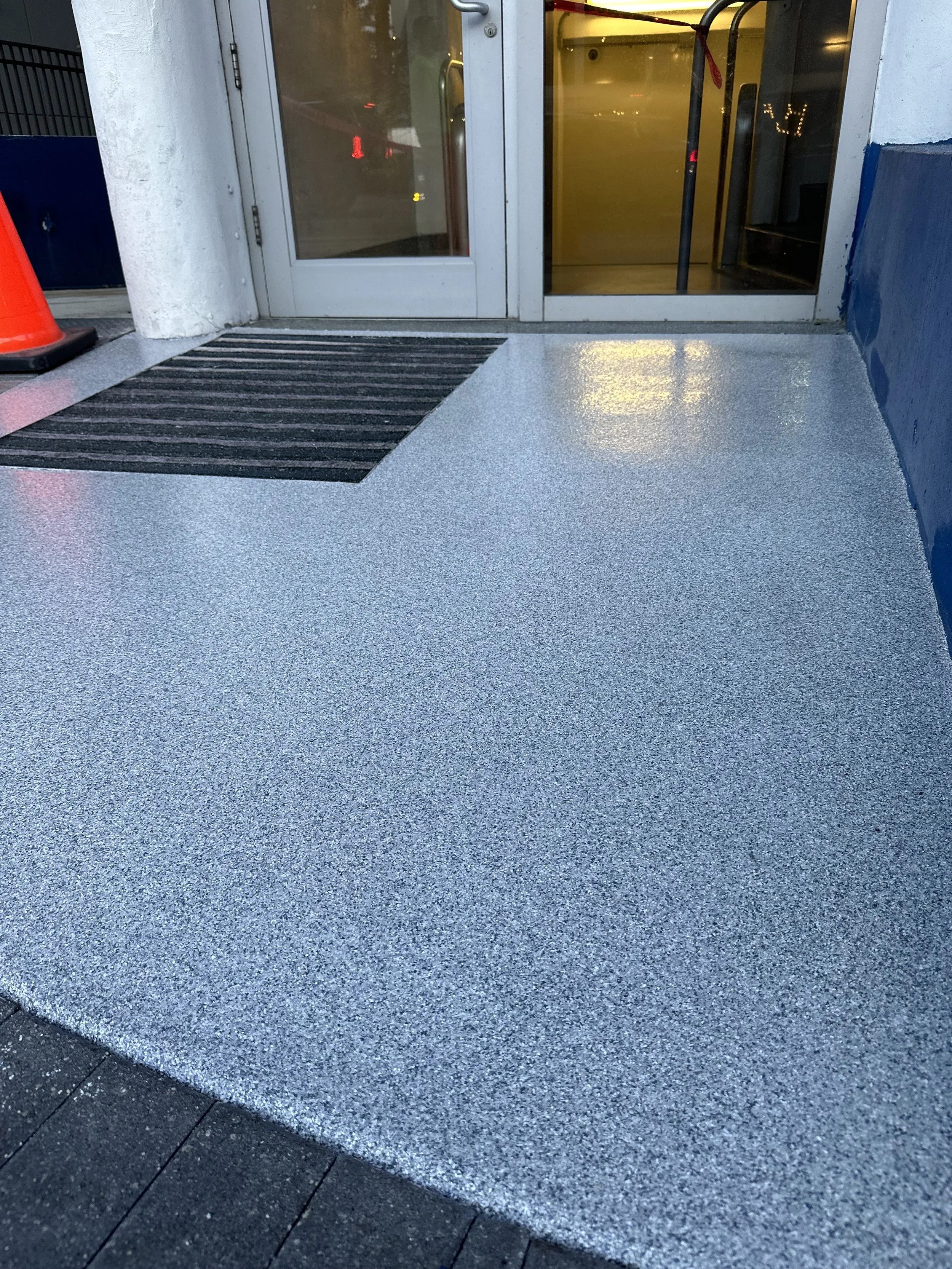 Outside entrance of a building with a glass door, a black welcome mat, and a textured grey and black sidewalk.