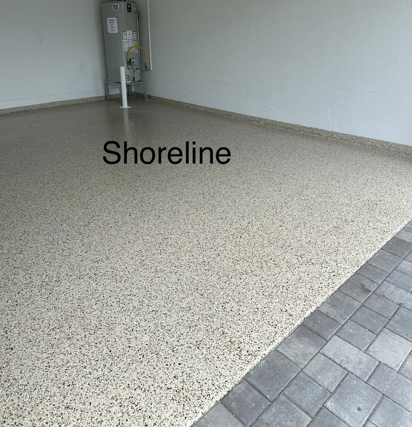 Empty area with a textured beige floor, the word 'Shoreline' overlaid, and a section of gray paving stones at the edge. In the background, there's a utility water heater against a white wall.