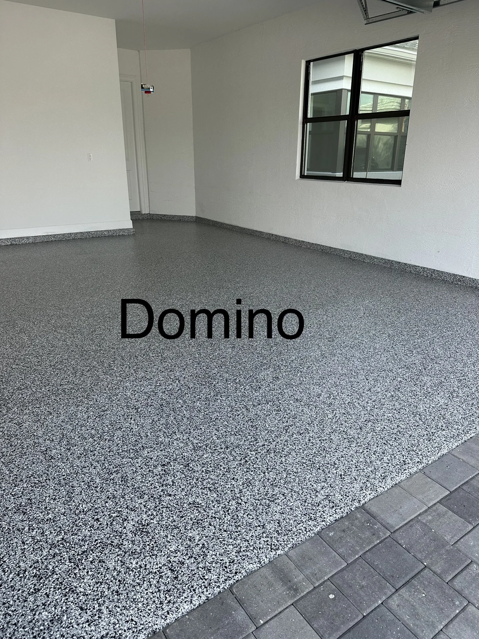 Interior of a house with freshly finished speckled epoxy-coated garage floor, labeled 'Domino', with a large window, white walls, and an open doorway.