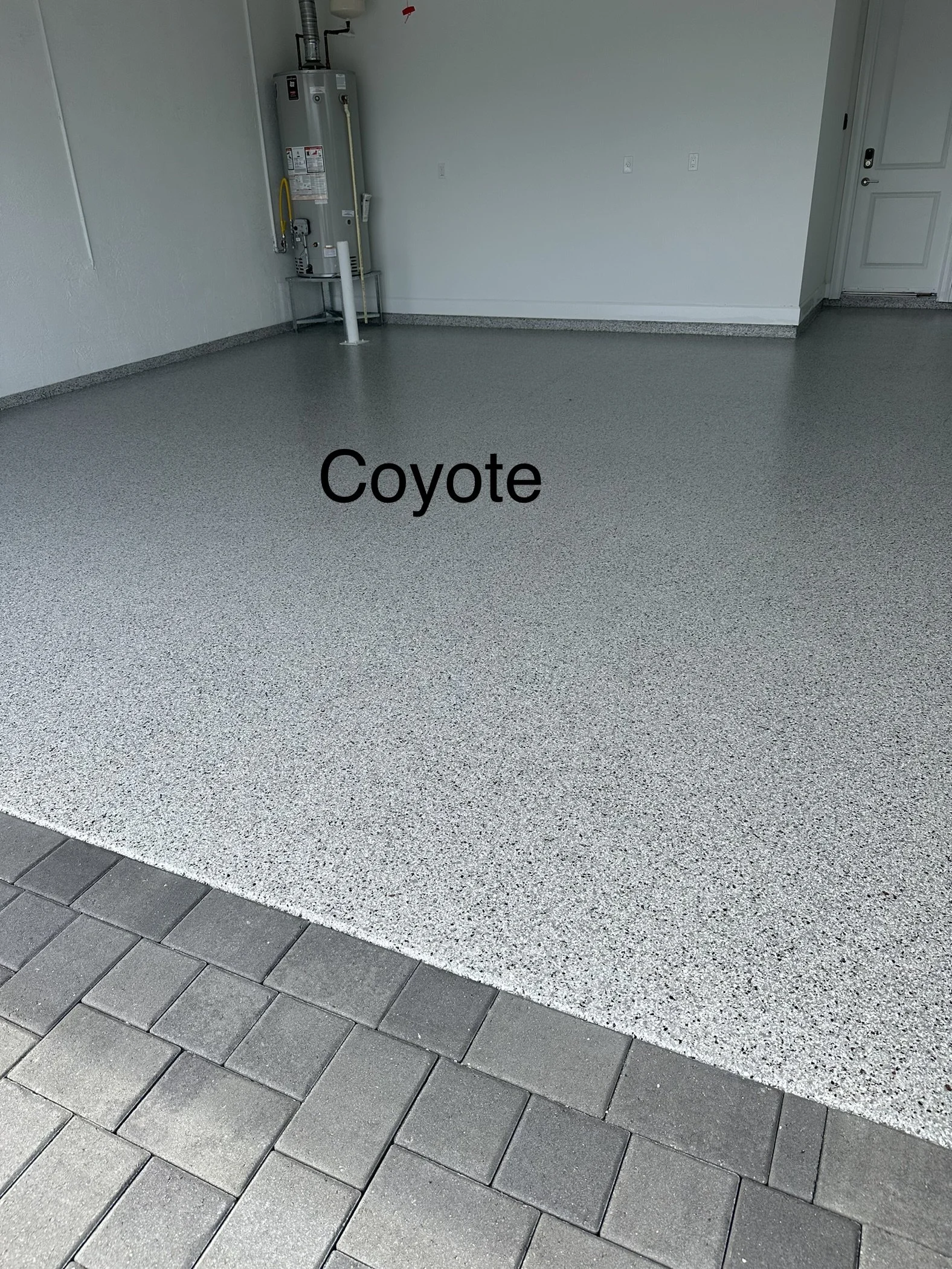 Garage with a newly coated floor labeled 'Coyote' with gray and black speckled epoxy, concrete pavers outside, water heater and utility hookups in corner.