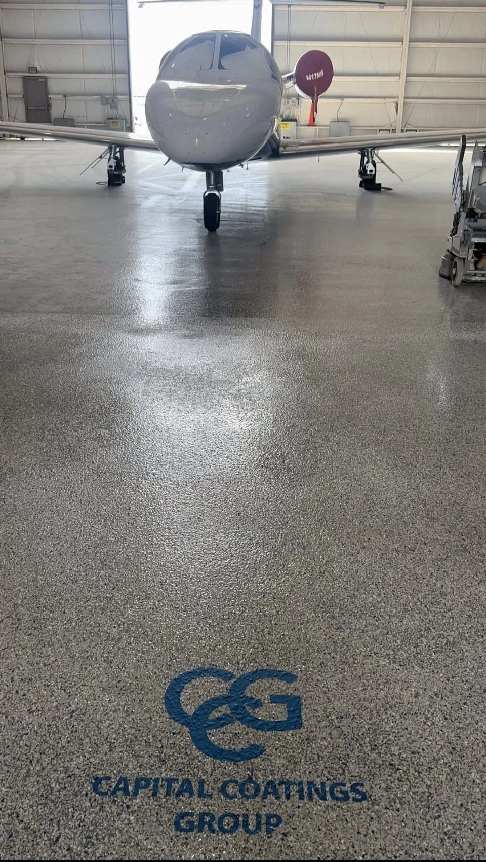 An airplane inside a hangar with a painted logo on the polished floor that says 'Capital Coatings Group' and a large fuel tank in the background.